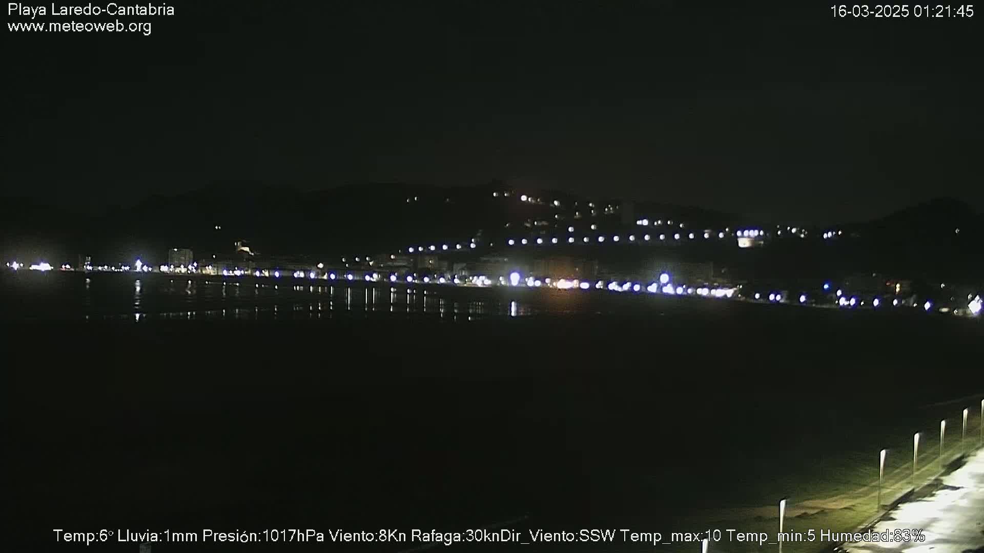 A nighttime view of a coastal town with lights reflecting on a calm body of water under a clear, dark sky.