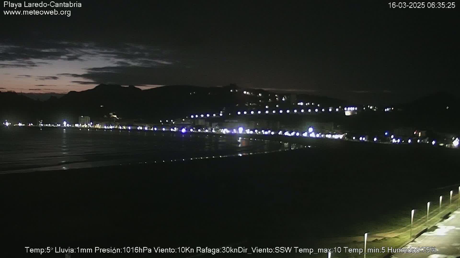A dark beach at night, lit by streetlights along a distant shoreline under a mostly clear, dark sky.