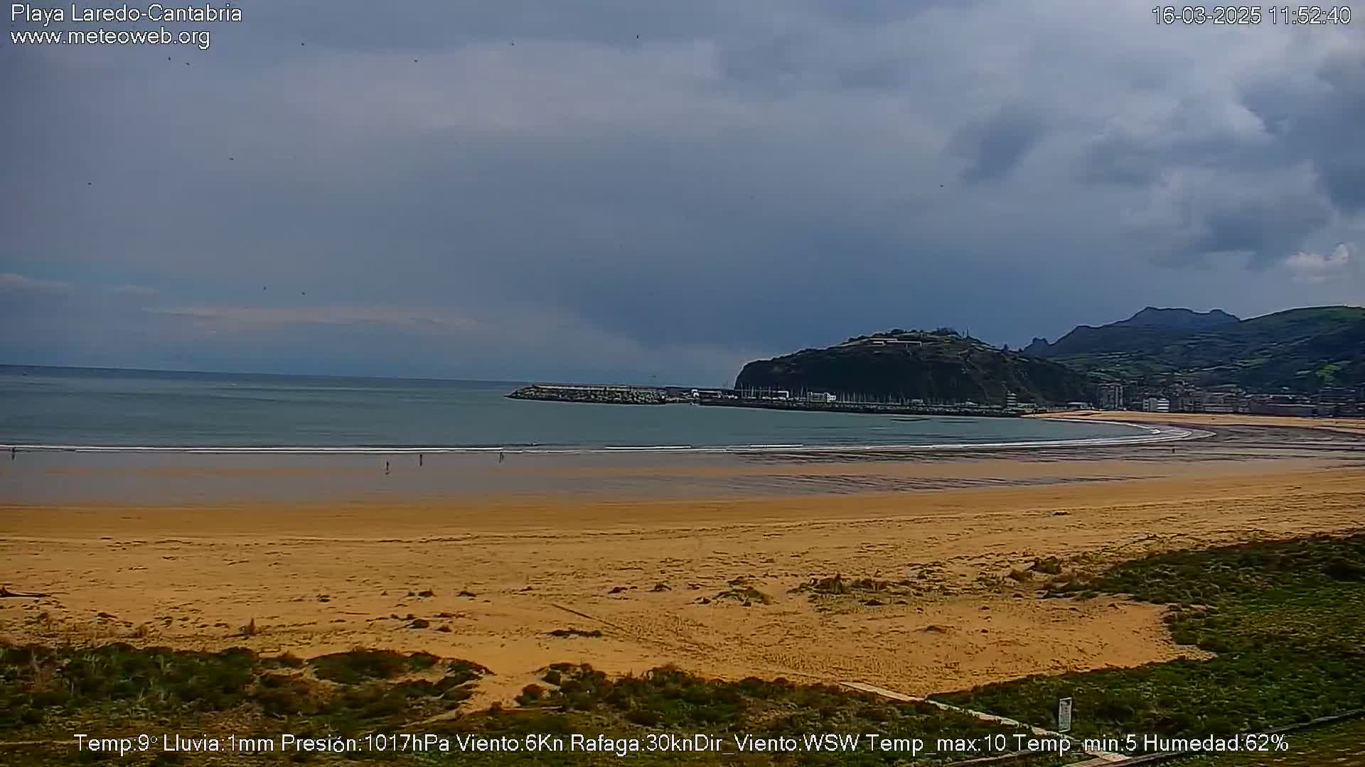 A sandy beach curves gently around a calm bay, backed by a hilly town under a mostly cloudy sky.