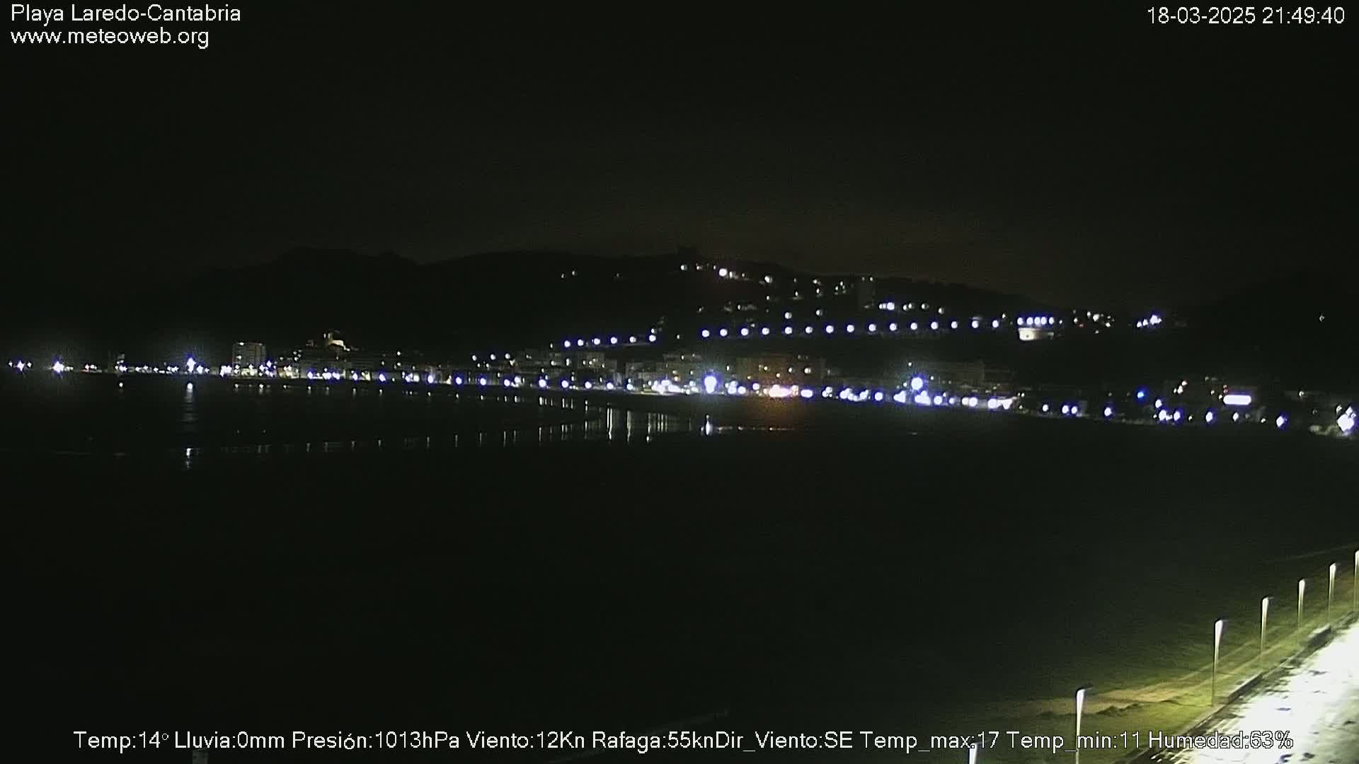 A nighttime view of a town's lights along a dark beach and hills under a clear sky.