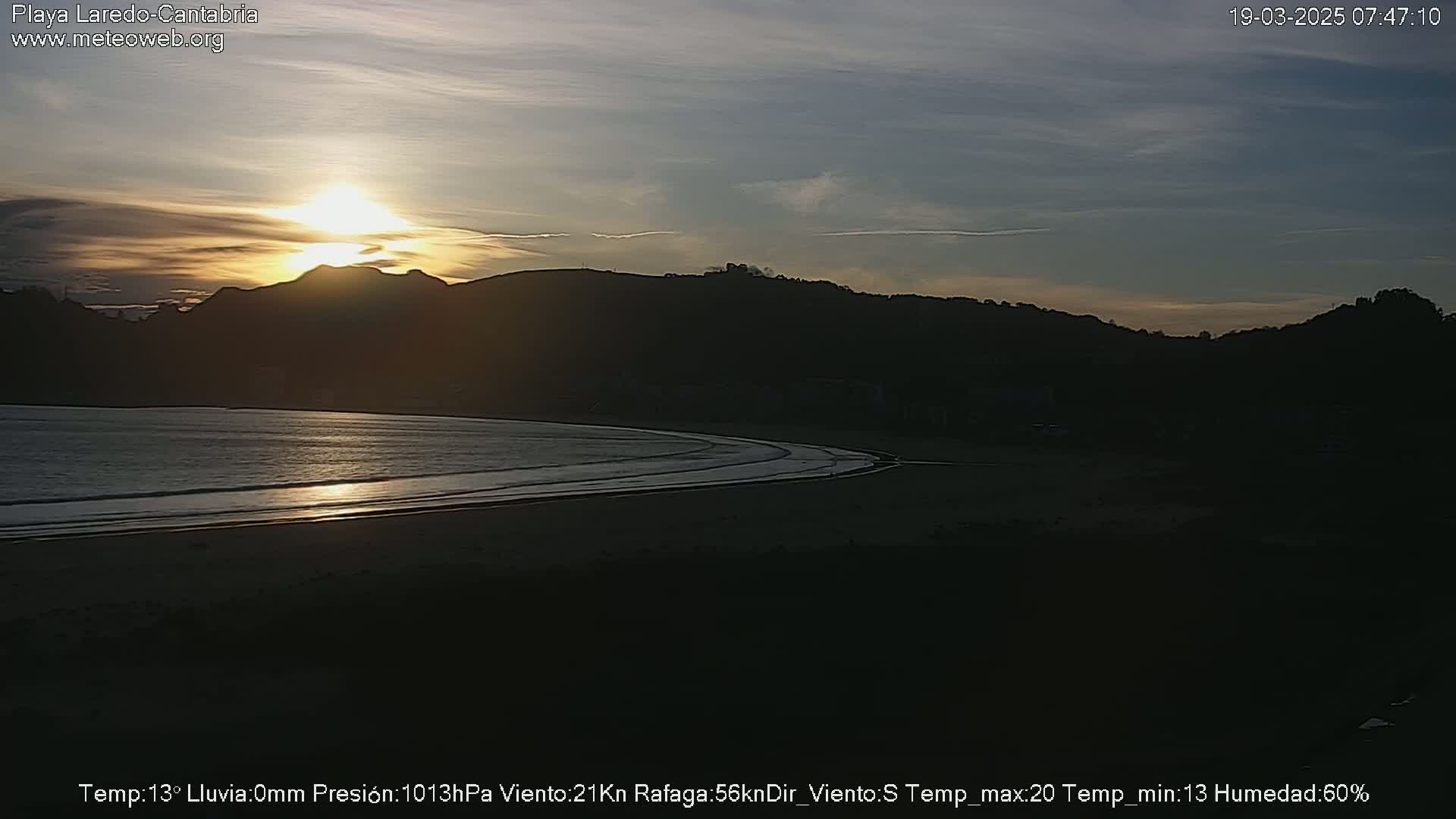 A sunset over a dark, sandy beach with calm waves, backed by silhouetted hills under a mostly clear sky.