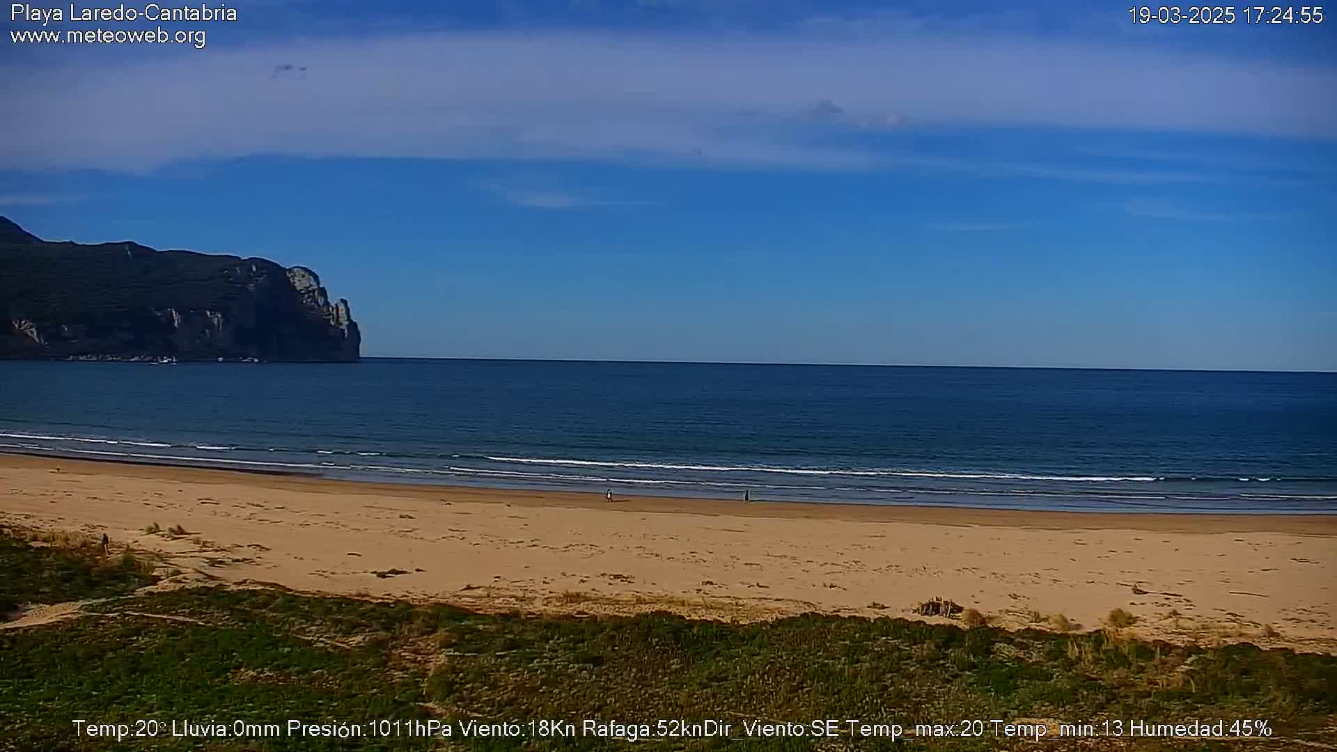 A sandy beach with calm ocean waves and a distant cliff face under a clear blue sky.