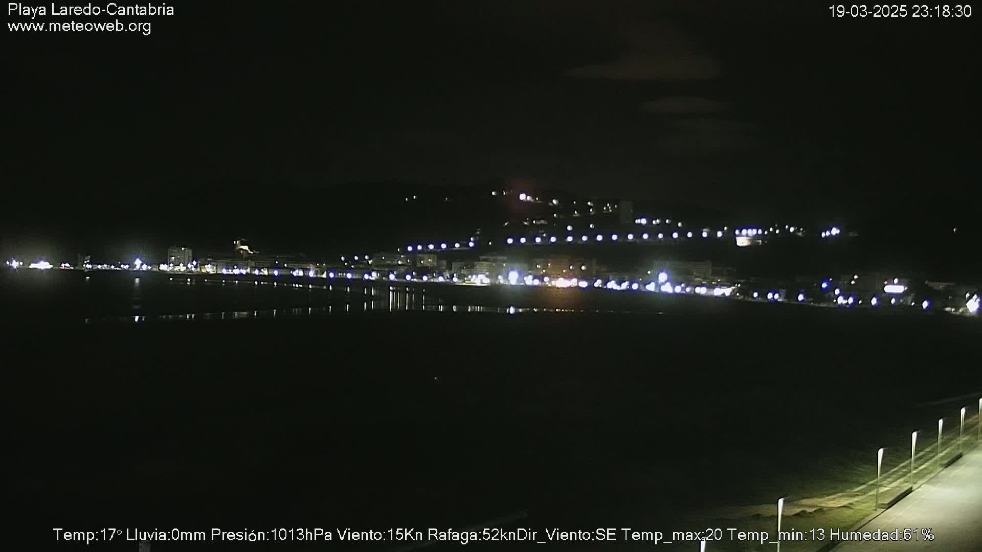 A nighttime view of a dark, calm beach with a town's lights illuminating the shoreline in the distance.