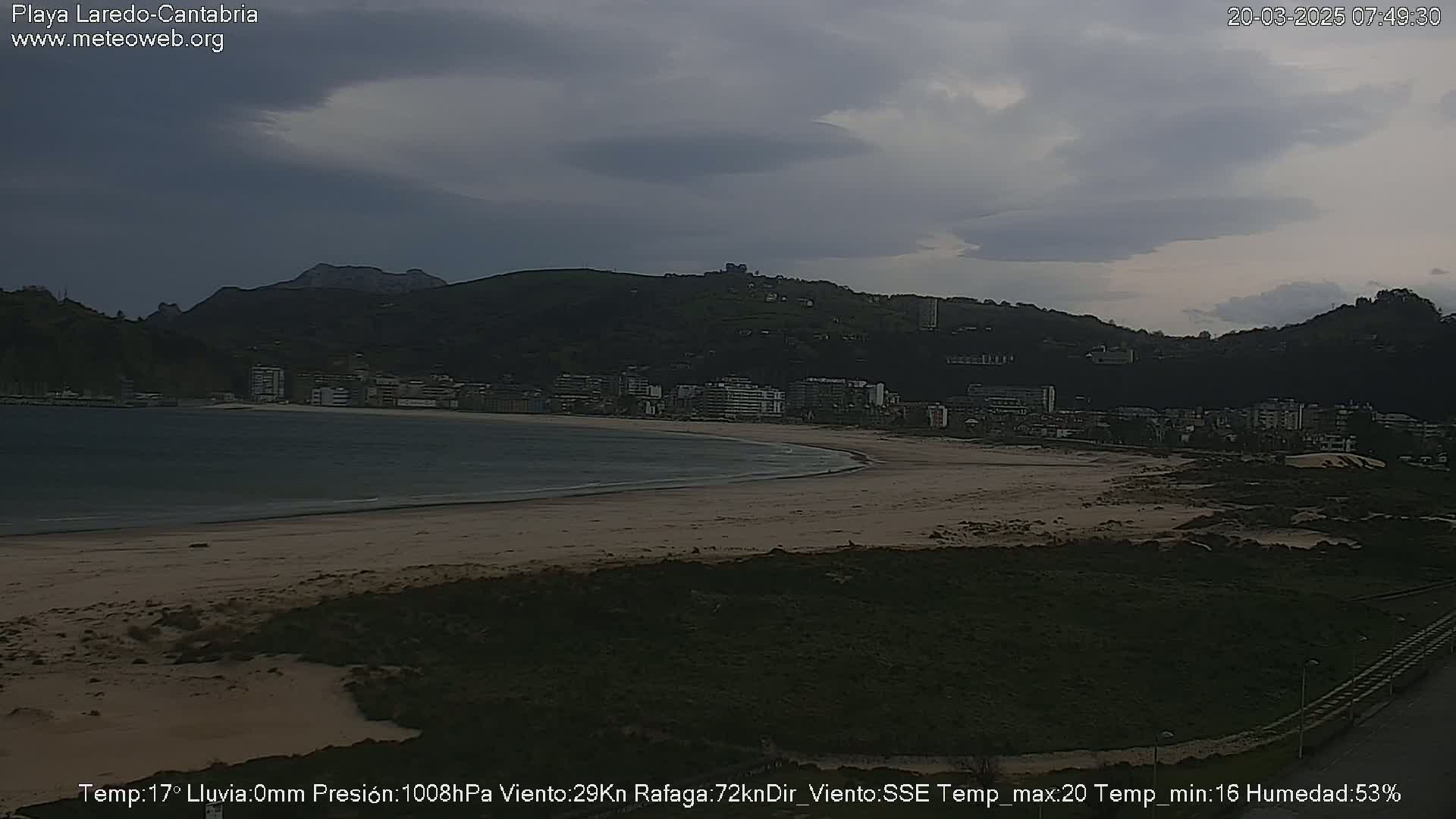 A sandy beach stretches along a calm bay, backed by a town nestled at the base of a dark, overcast hill.