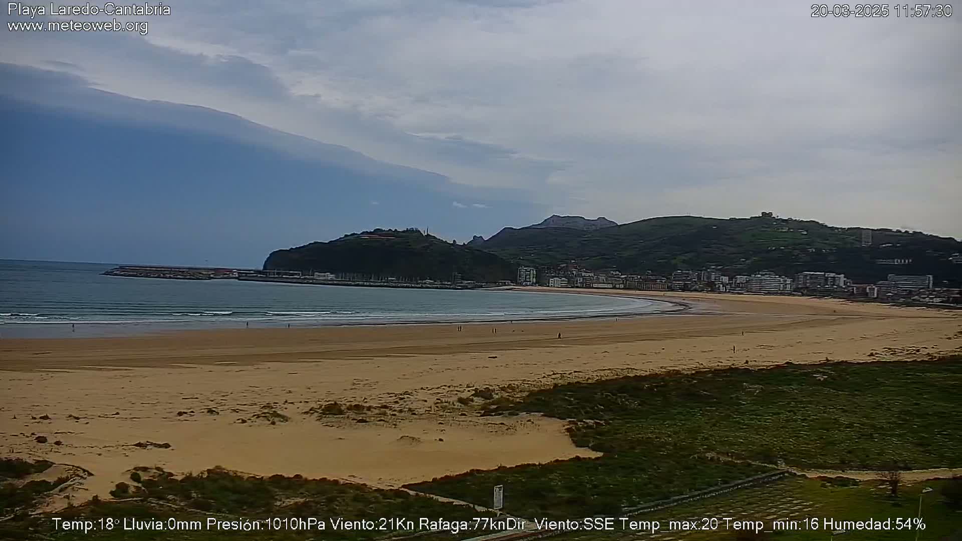 A sandy beach curves gently around a bay, backed by a town nestled against green hills under a mostly cloudy sky.