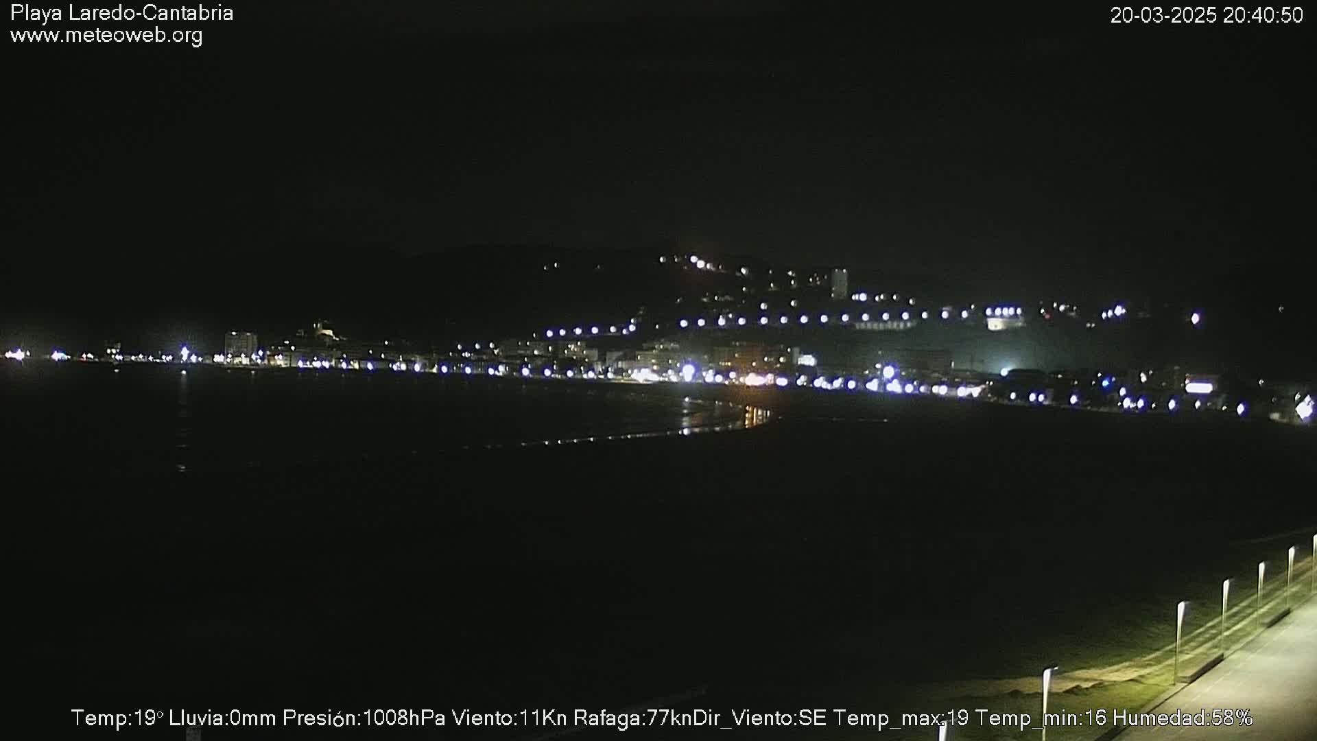 A nighttime view of a coastal town with lights illuminating buildings and a dark beach under a clear sky.