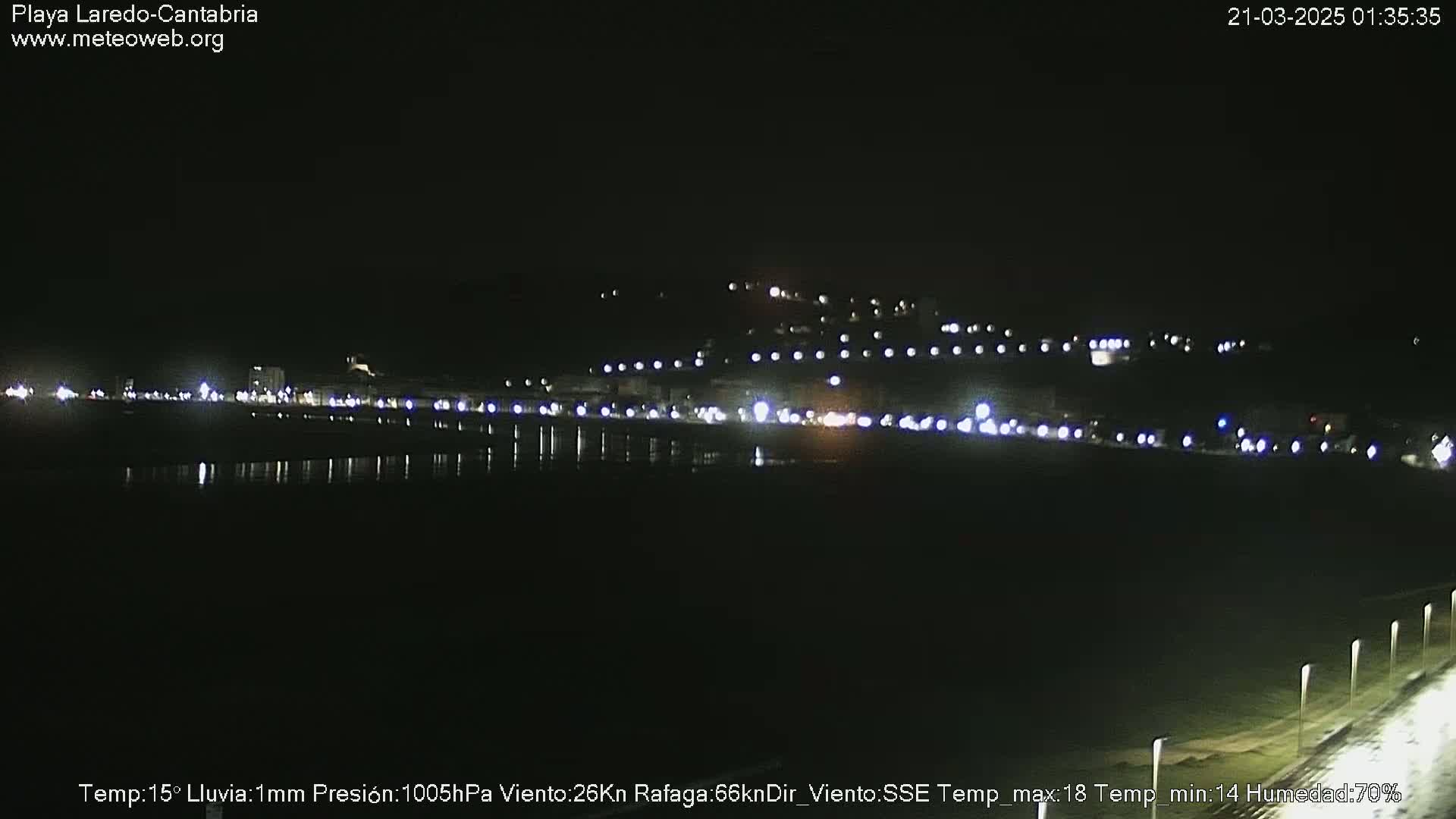 A nighttime view of a coastal town with lights reflecting on calm water, under slightly rainy conditions.