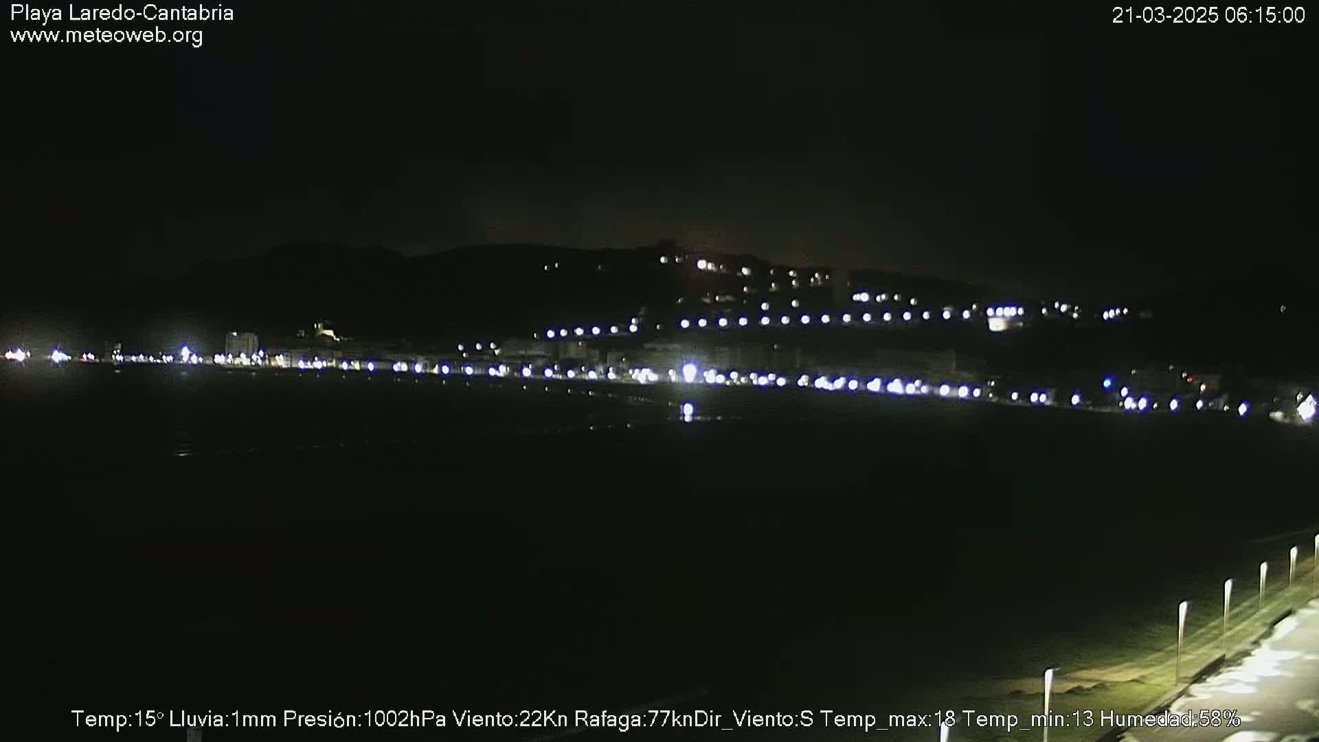 A nighttime, dimly lit coastal town is seen under light rain, with a dark hill in the background.