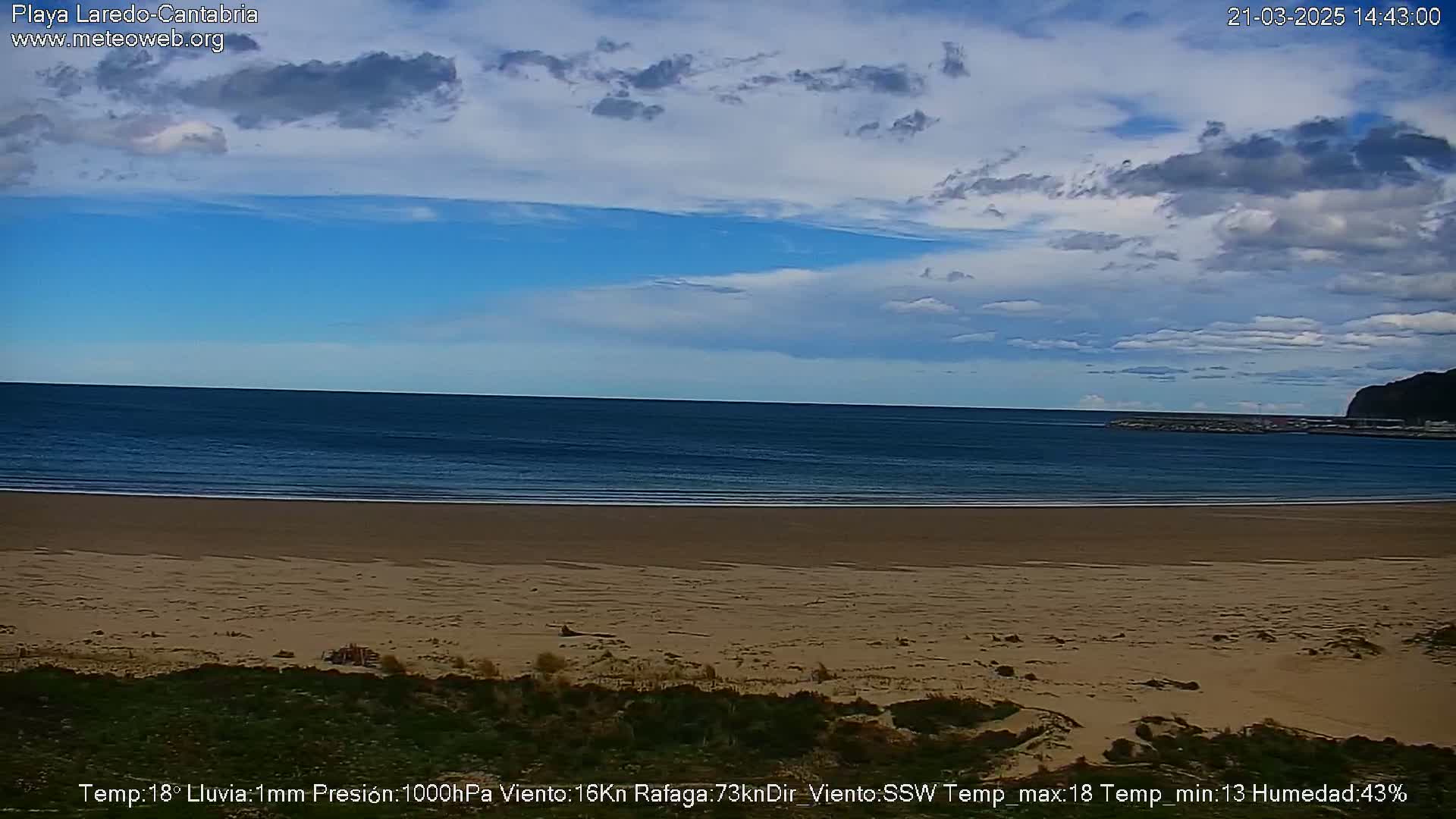 A sandy beach stretches to a calm ocean under a partly cloudy blue sky.