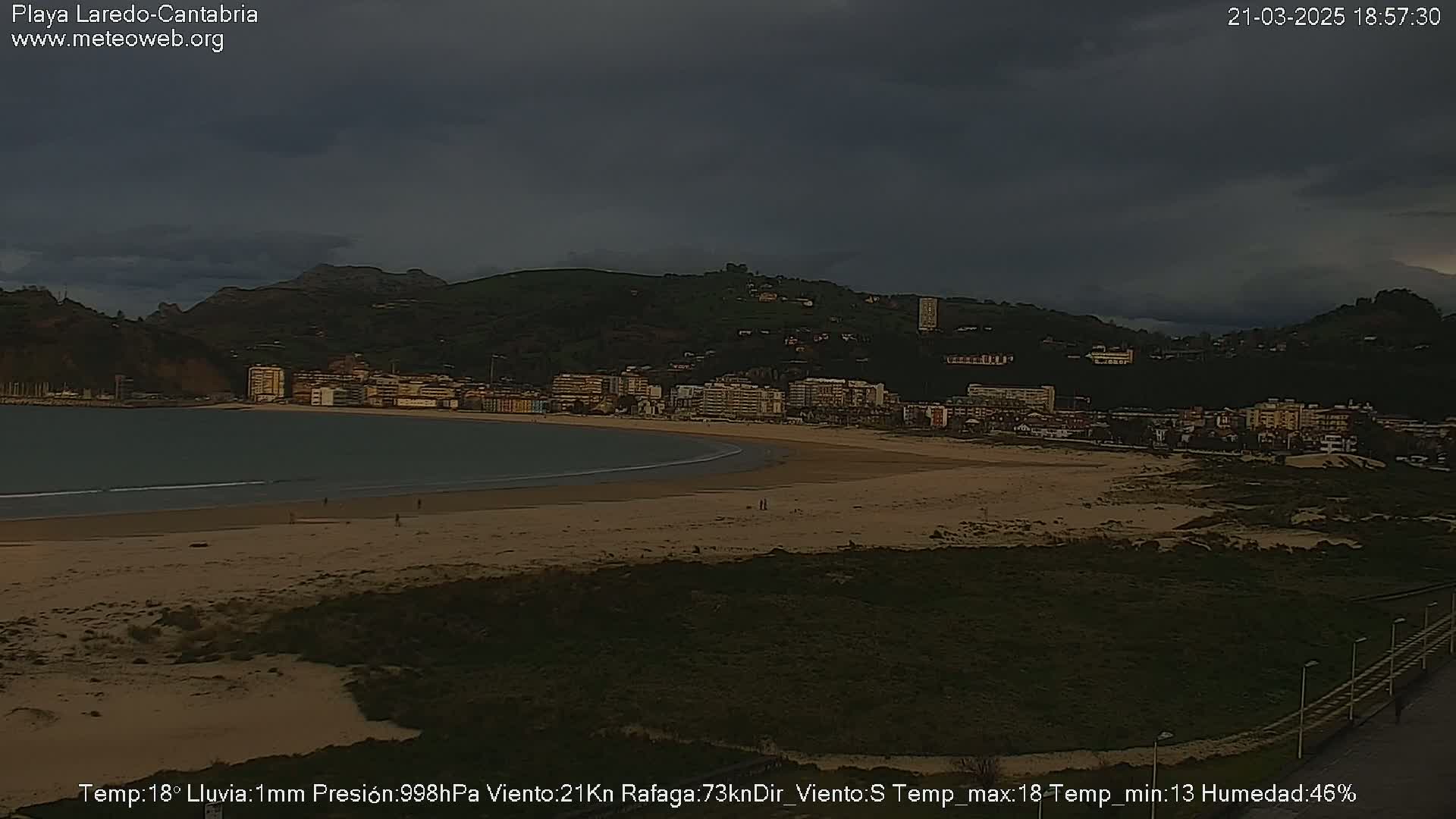 A sandy beach with a town and hills in the background under a dark, cloudy sky with light rain.