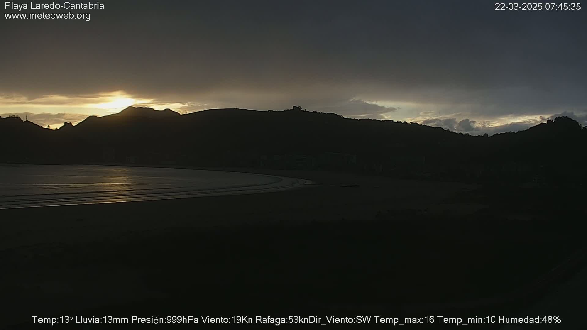 A dark beach at sunset, with a low-lying hill silhouetted against a partly cloudy sky.