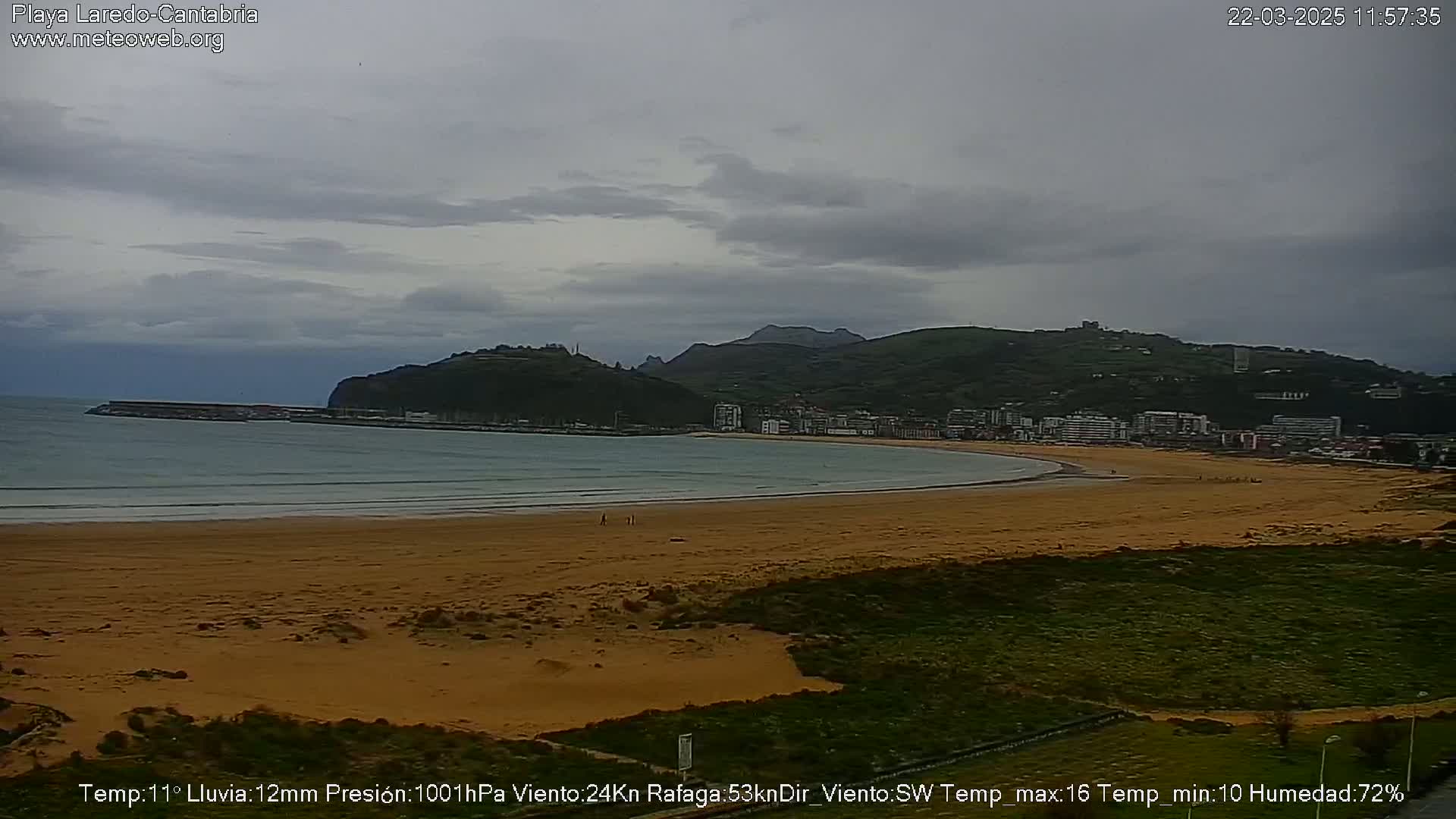 A sandy beach stretches below a cloudy sky, with a town and hills visible in the distance, and light rain is falling.