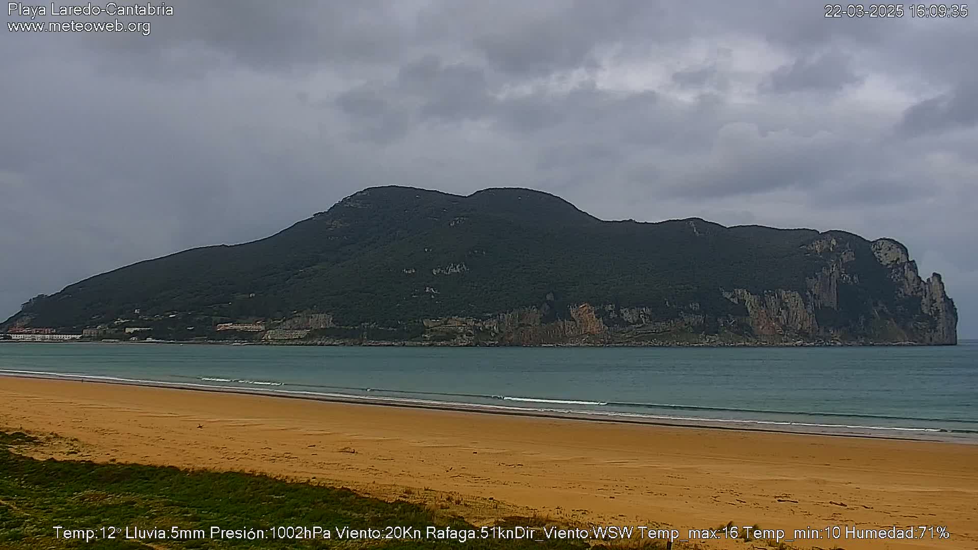 A sandy beach stretches towards a large, dark-green mountain rising from the calm, teal ocean under a mostly cloudy sky.