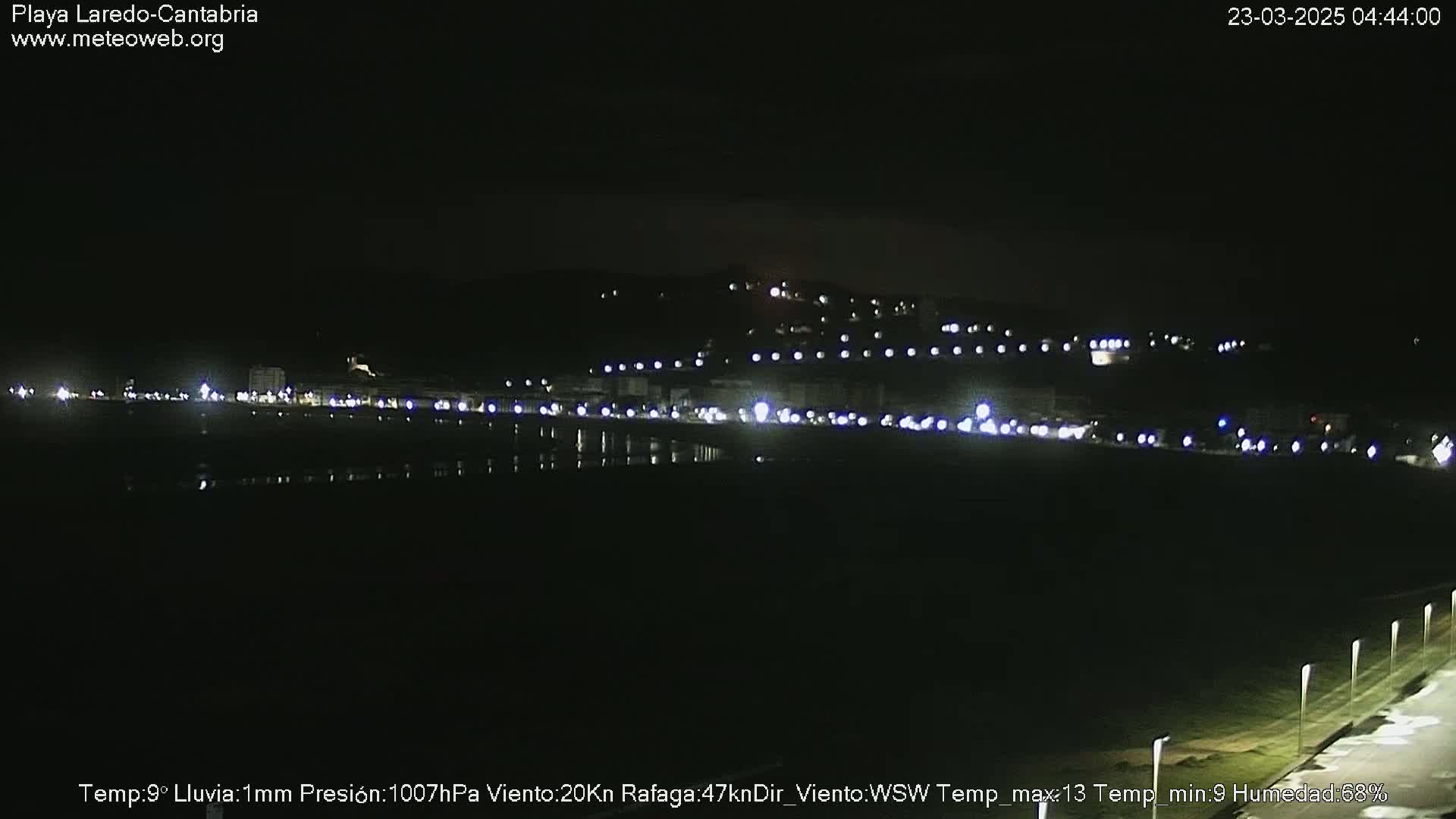 A nighttime view of a dark, calm body of water reflecting the lights of a coastal town under a clear sky.