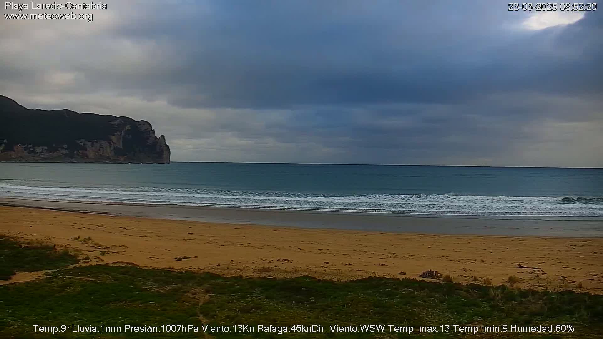 A sandy beach with gentle waves laps the shore, under a mostly cloudy sky, with a large rocky cliff visible in the distance.