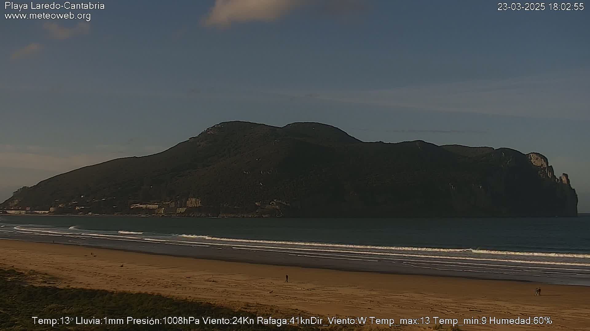 A sandy beach with gentle waves laps against the shore, under a mostly clear sky, with a large, dark, mountainous landmass visible in the background.