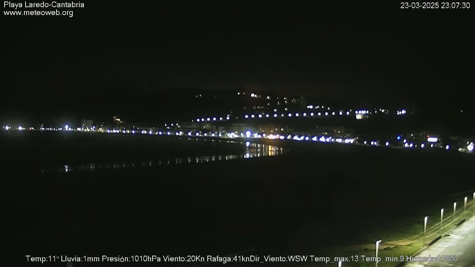 A nighttime view of a dimly lit beach town, with faint lights reflecting on the water under a dark sky with light rain.