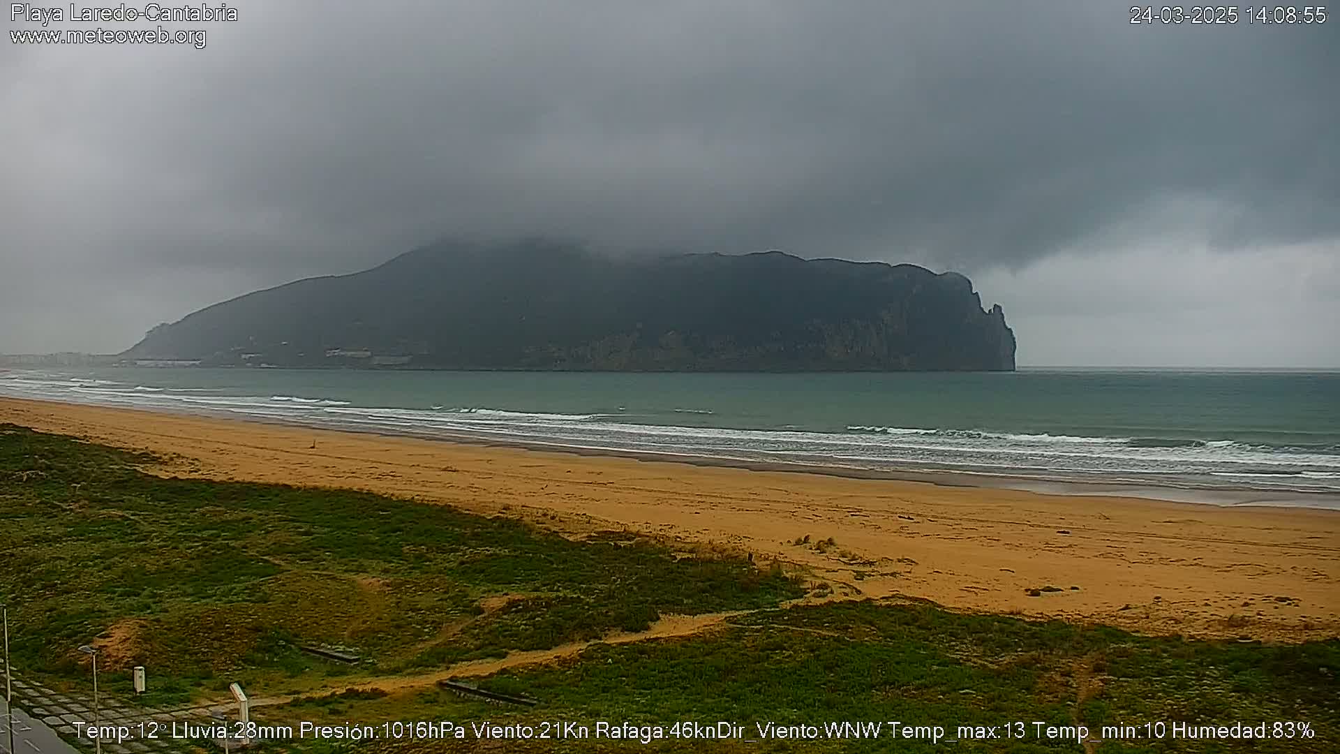 A sandy beach with gentle waves stretches towards a dark, mountainous headland under a cloudy, overcast sky.