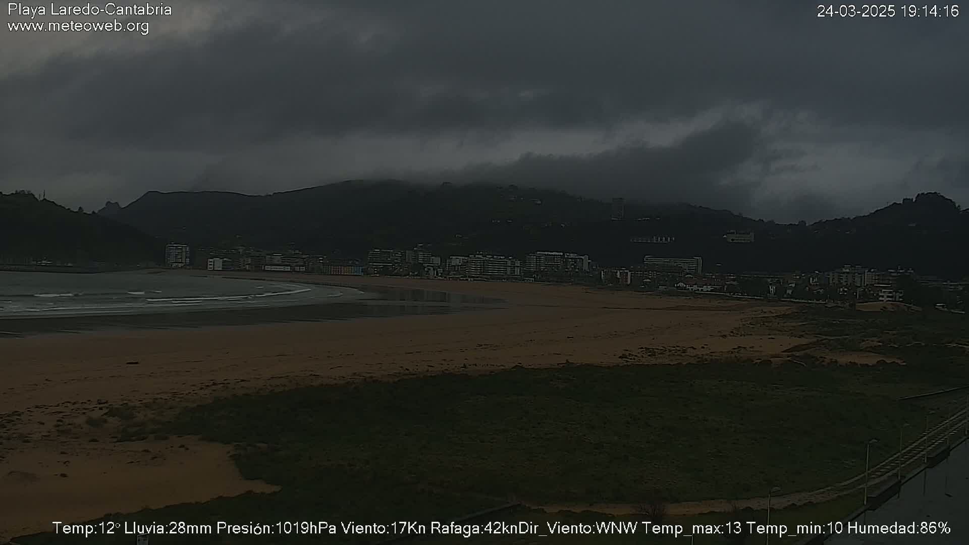 A dark, overcast beach scene shows a town nestled against hills under a gray sky, with a calm ocean and sandy beach in the foreground.