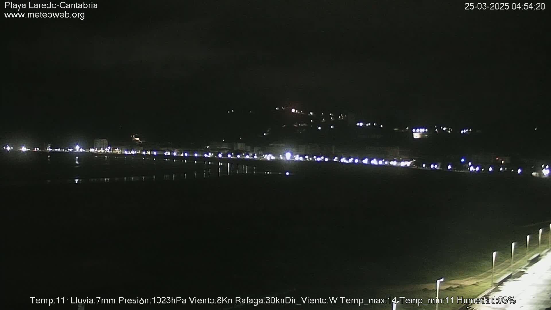 A nighttime view of a coastal town illuminated by streetlights, with rain having recently fallen.