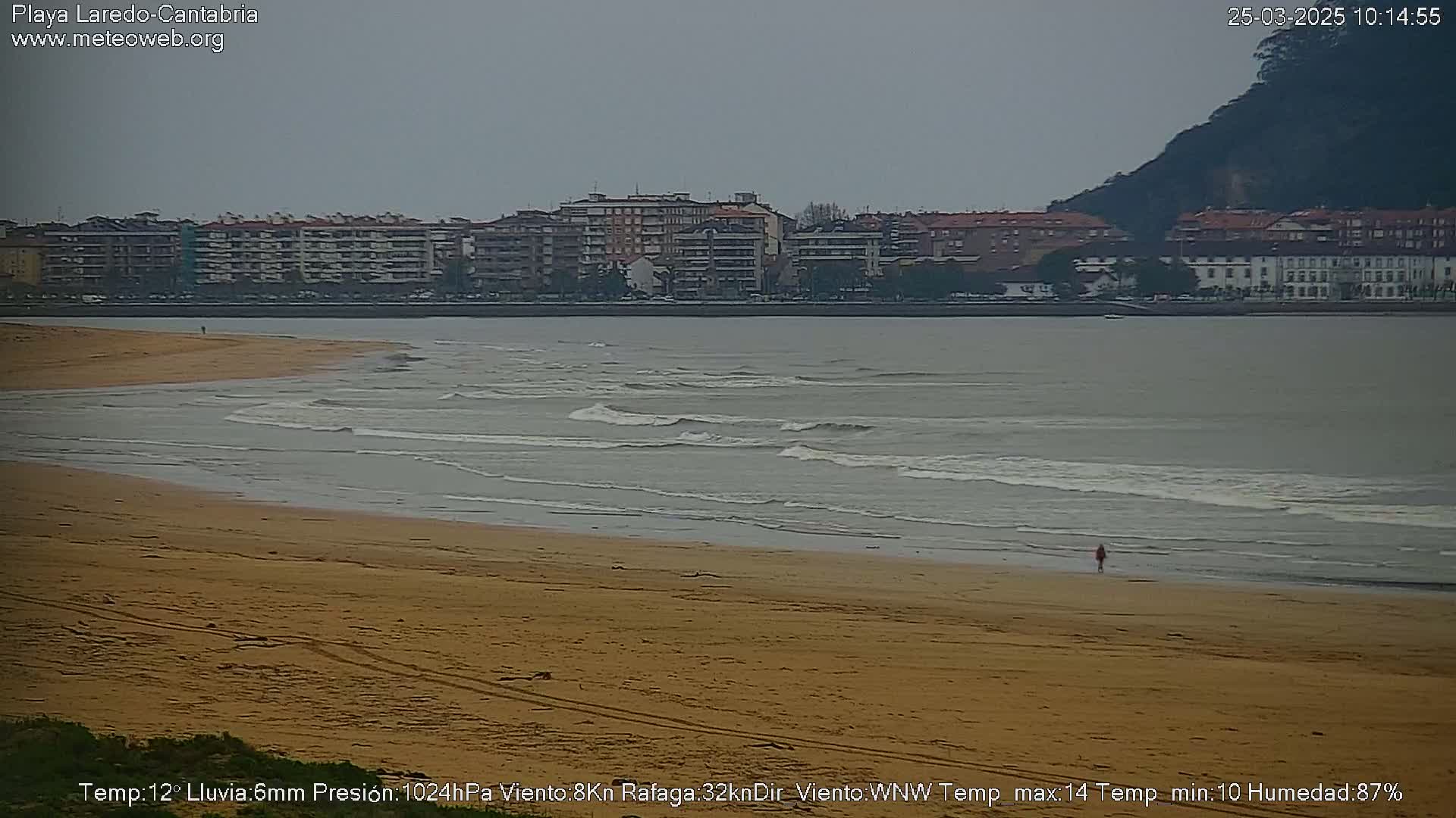 A person walks on a sandy beach with small waves lapping the shore, under an overcast sky, in front of a town situated next to a hill.