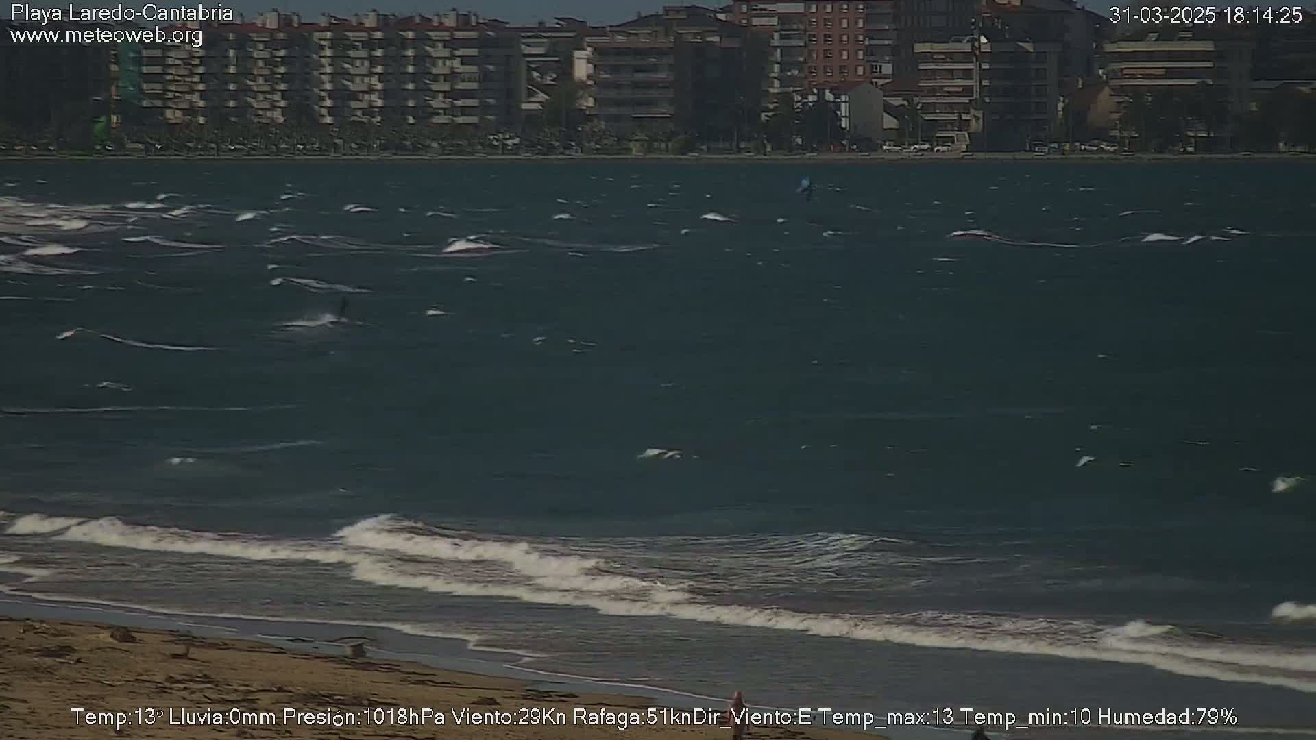 A windy, choppy ocean with waves breaking on a sandy beach in front of a line of apartment buildings.