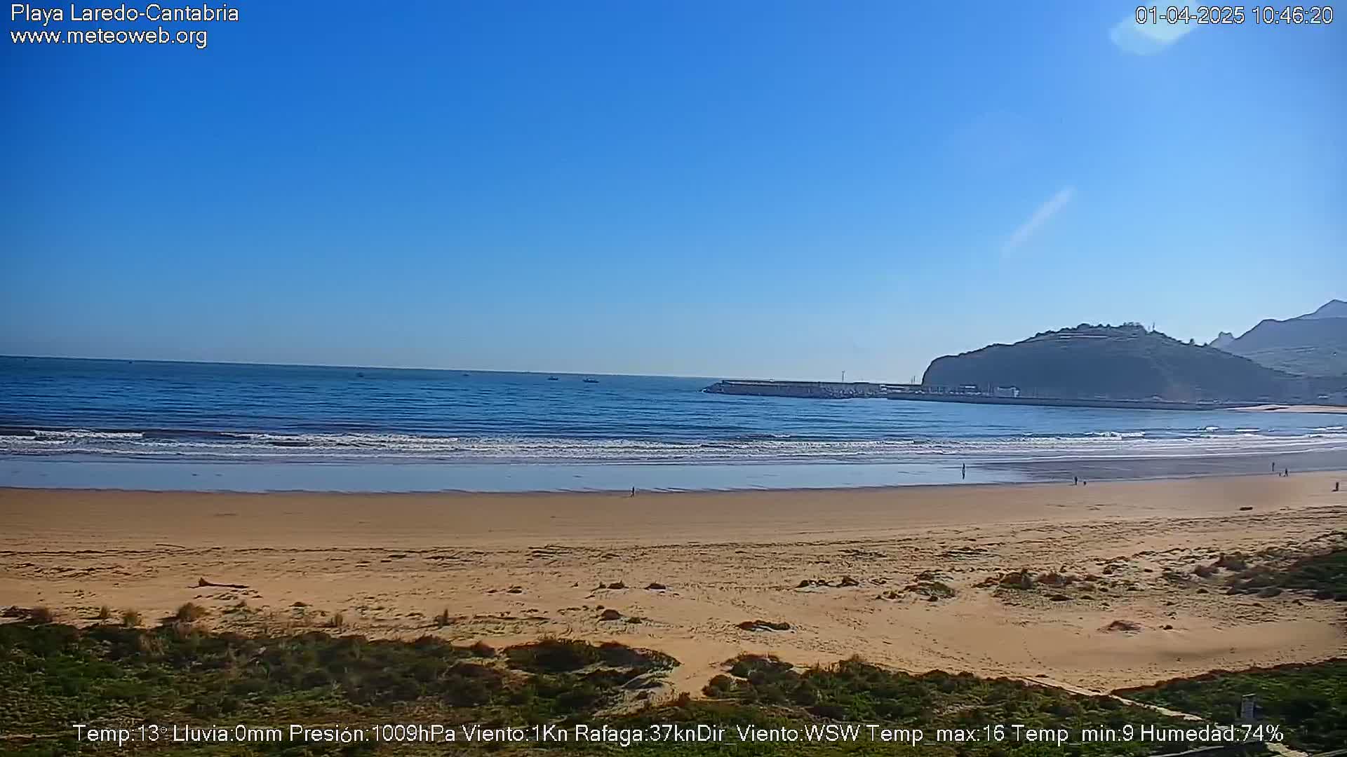 A sandy beach with gentle waves laps at the shore under a clear blue sky, a green hill rising in the background.