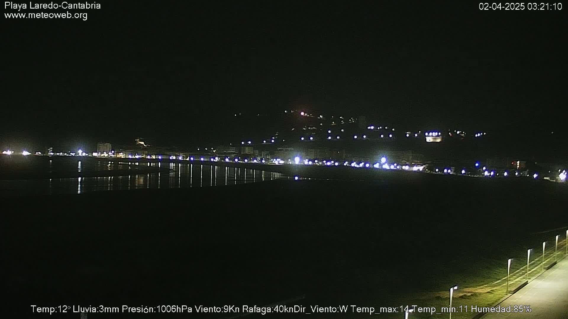 A nighttime view of a coastal town's lights reflecting on calm water under a dark sky with light rain.