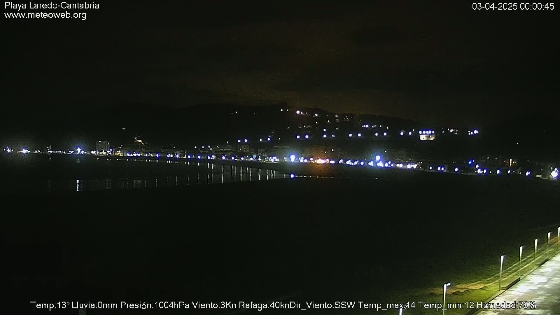 A nighttime view of a dark, calm beach with a line of lights from buildings on the distant hill.