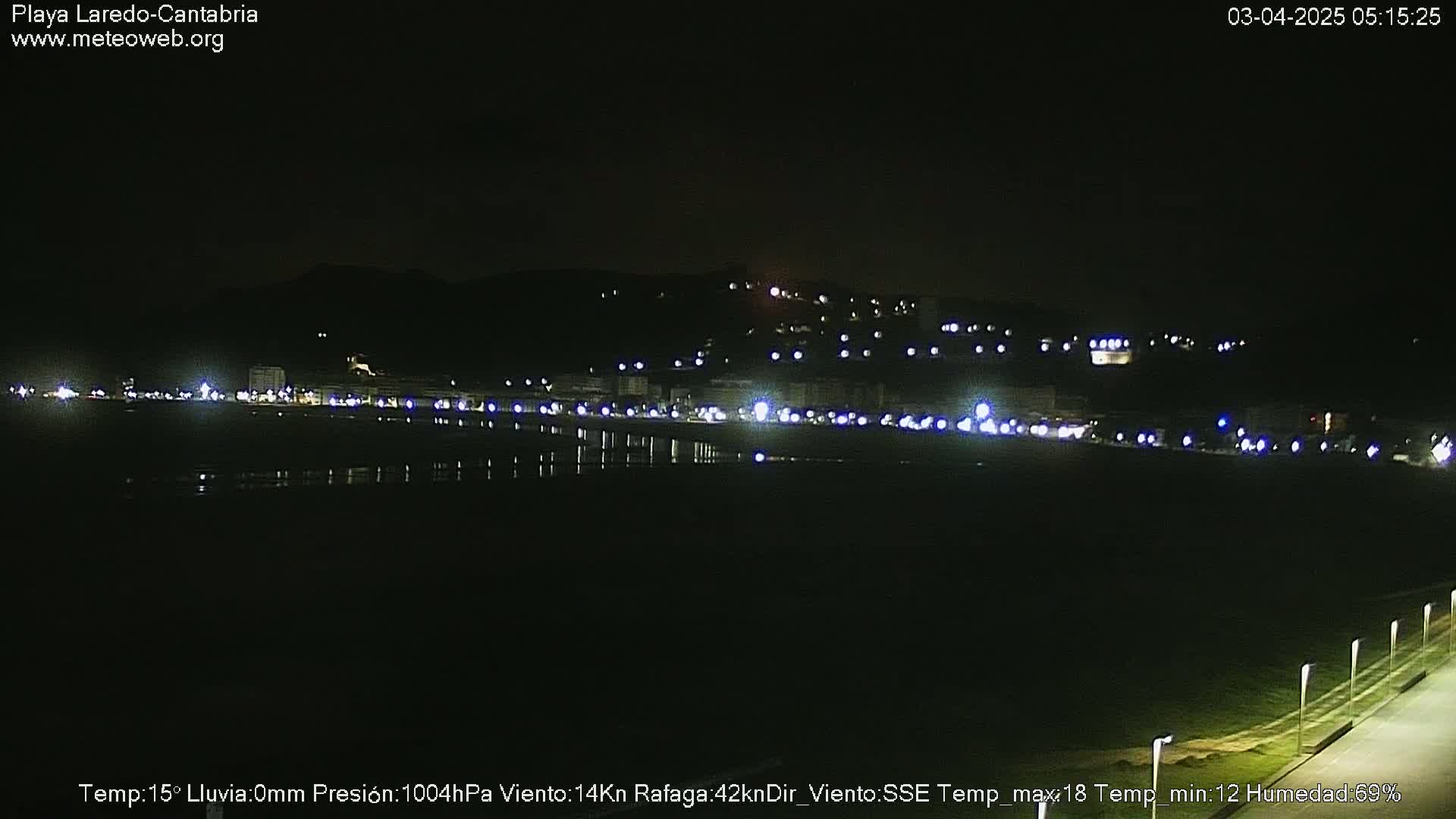 A nighttime view of a dark beach and a dimly lit town situated against a dark hillside under a clear sky.