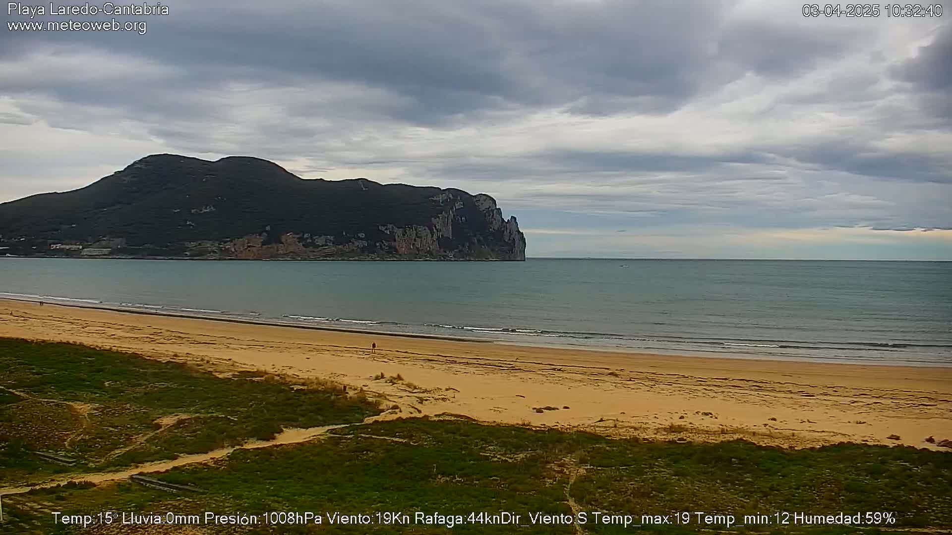 A sandy beach stretches before a calm sea, under a mostly cloudy sky, with a large, dark mountain in the background.