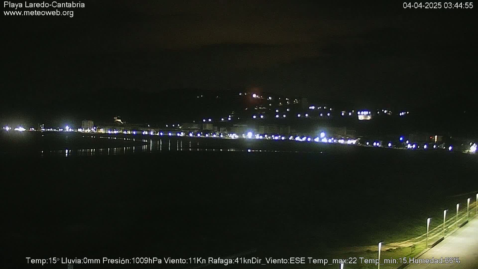 A nighttime view of a dimly lit beach and town, with a calm body of water reflecting the lights.