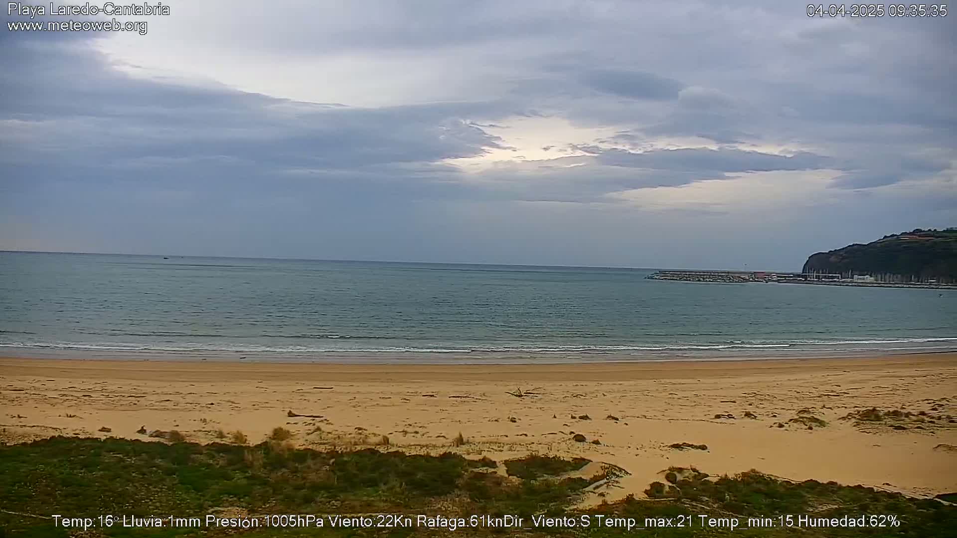 A sandy beach stretches towards a calm ocean under a mostly cloudy sky.