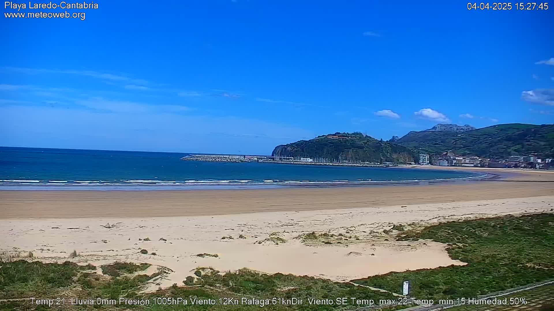 A sandy beach with calm waves and a clear blue sky stretches out to a bay, backed by green hills and a small town.