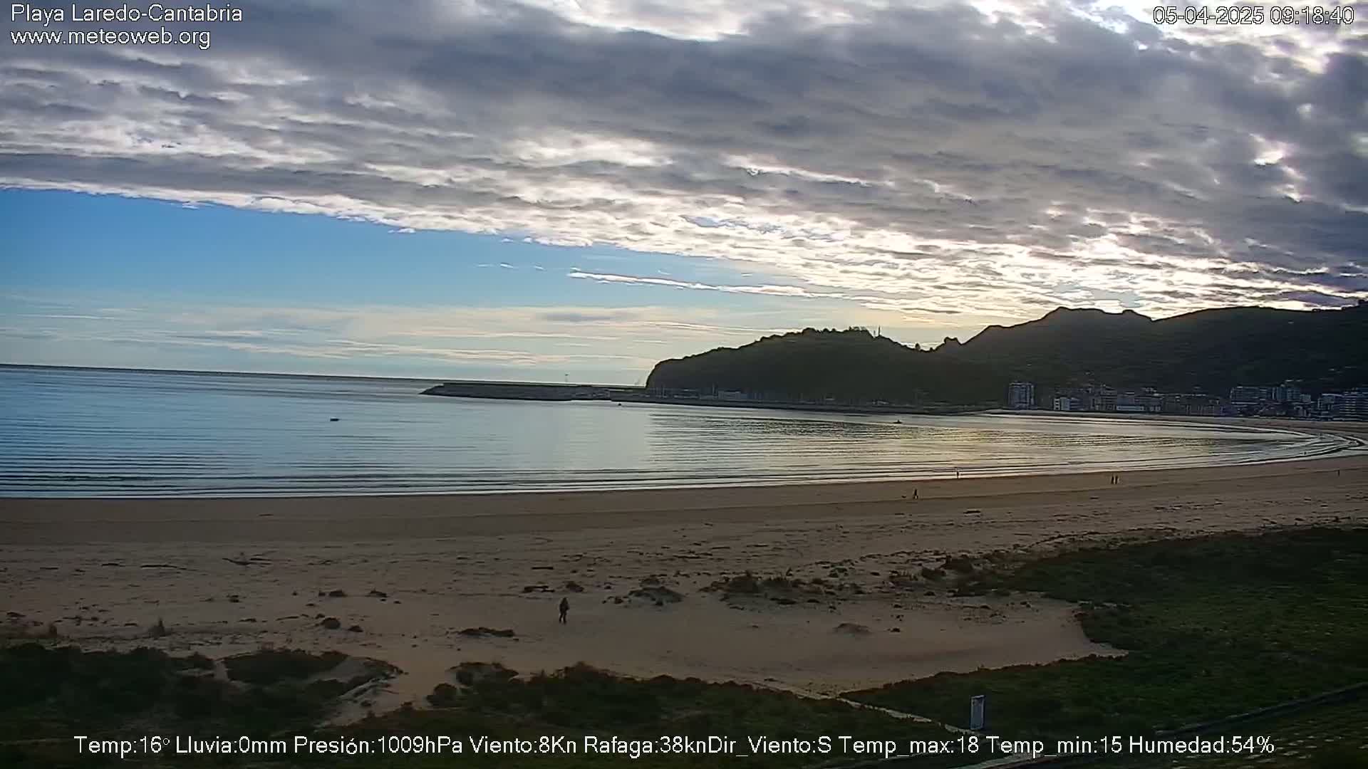 A mostly cloudy day shows a sandy beach curving around a calm bay, backed by a hill and town.