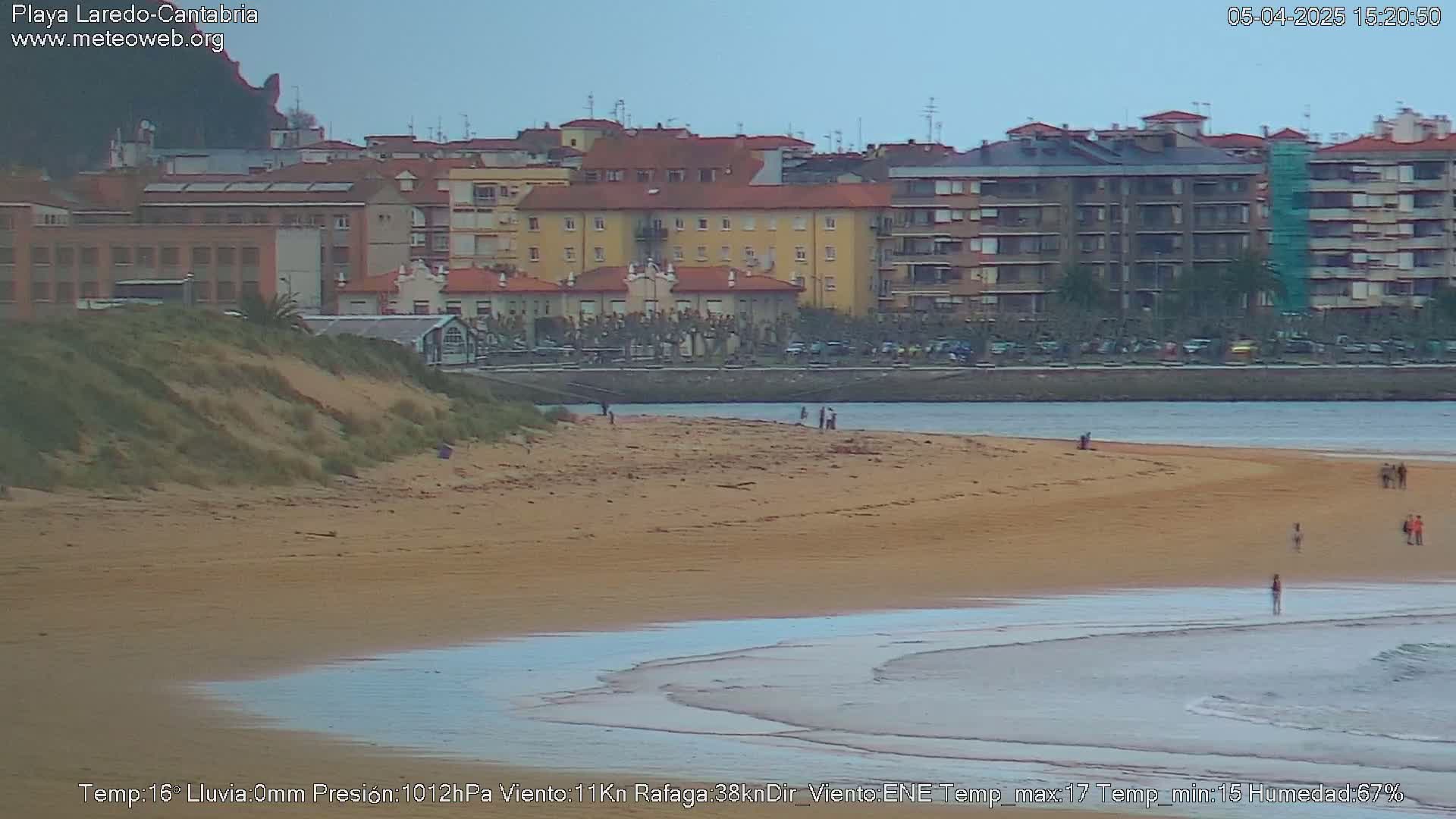 A sandy beach with a few people on it, next to buildings and a calm body of water under an overcast sky.