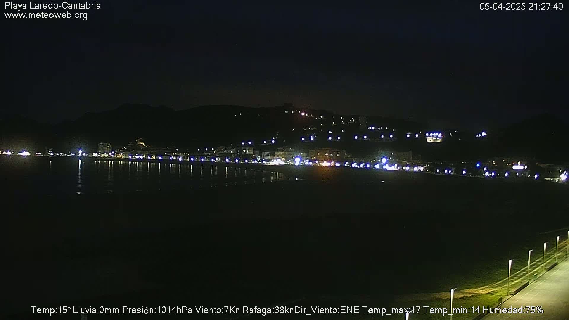 A nighttime view of a dark beach and city lights reflecting on calm water under a clear sky.