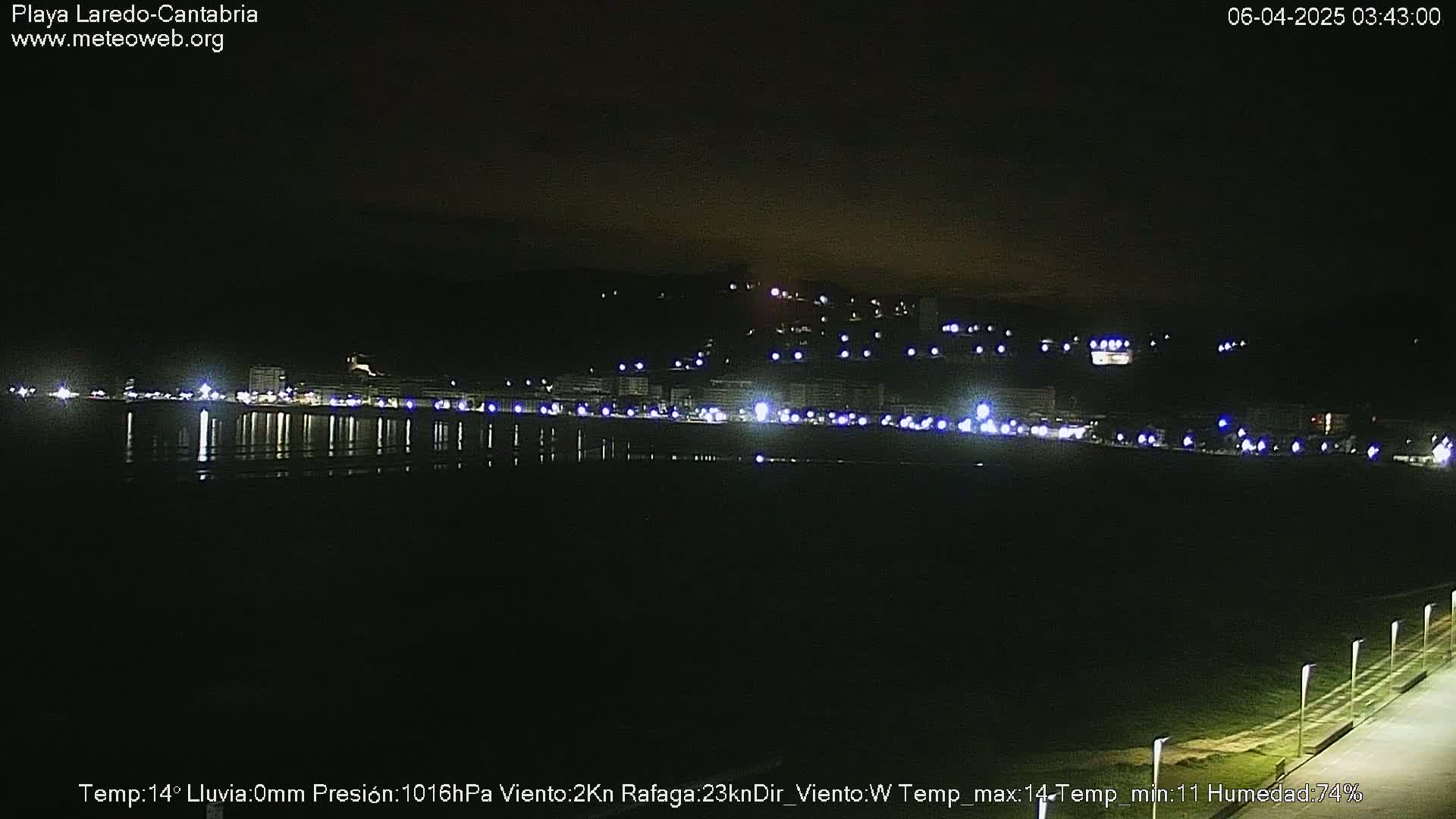 A nighttime view of a calm, dark beach with a town's lights reflected in the water.