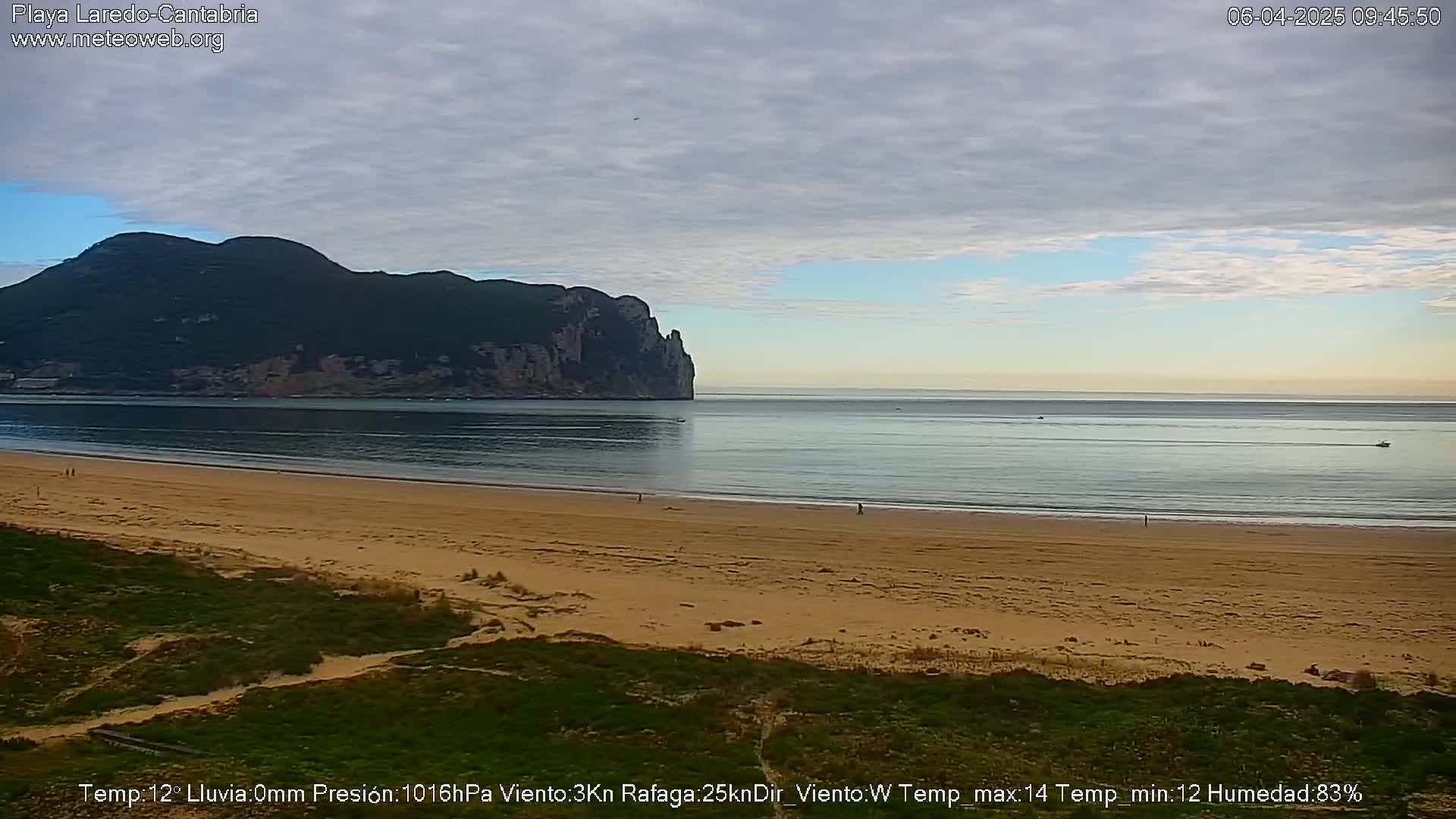 A mostly cloudy day shows a sandy beach, calm ocean, and a large, dark mountain in the distance.