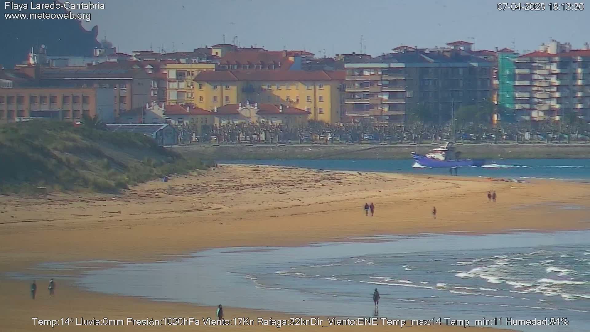 A sandy beach with a few people walking along the shore and a small boat in the calm water, backed by a town under a clear sky.