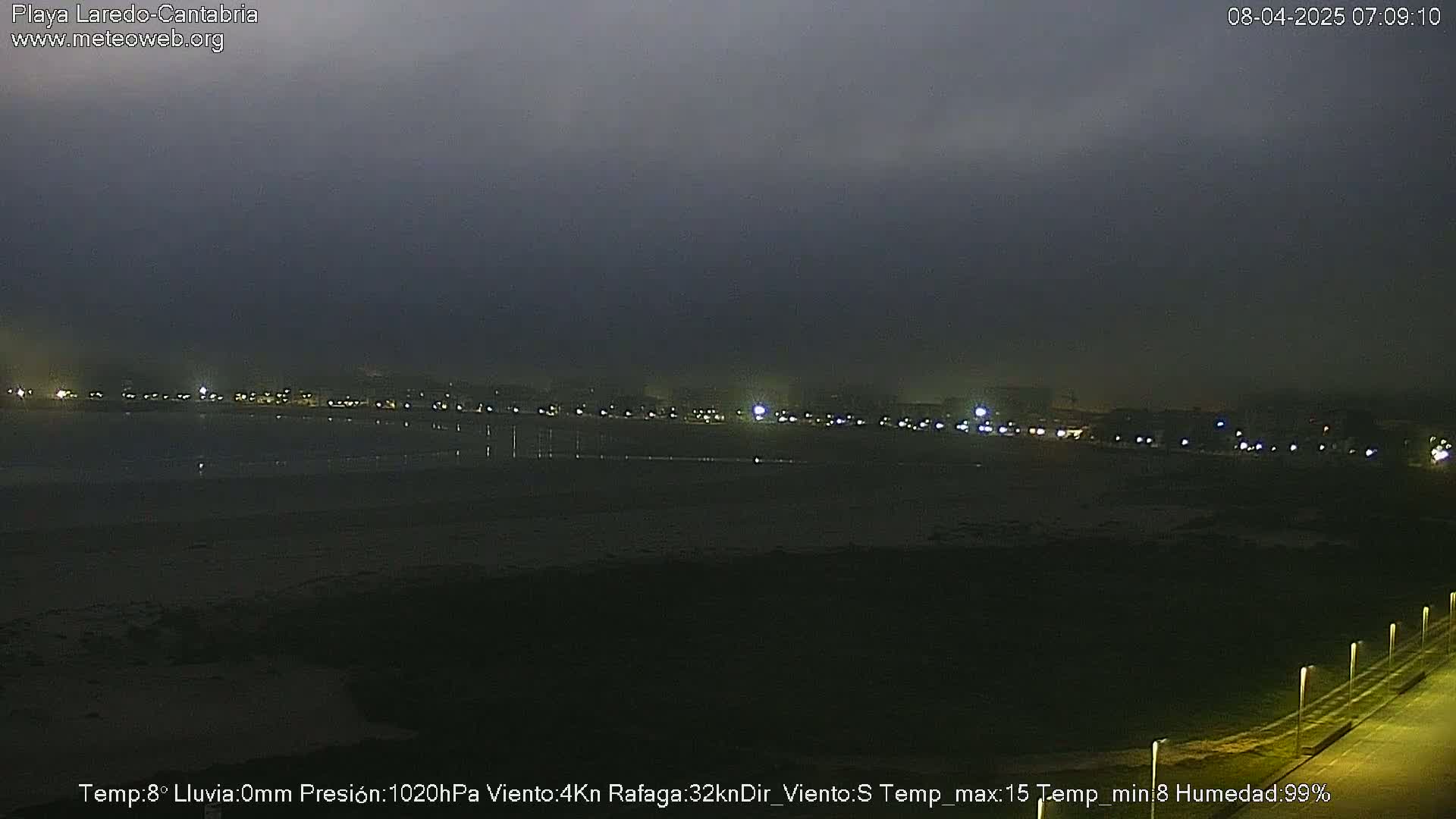 A dark, overcast night scene shows a dimly lit beach and shoreline with a line of lights in the distance.