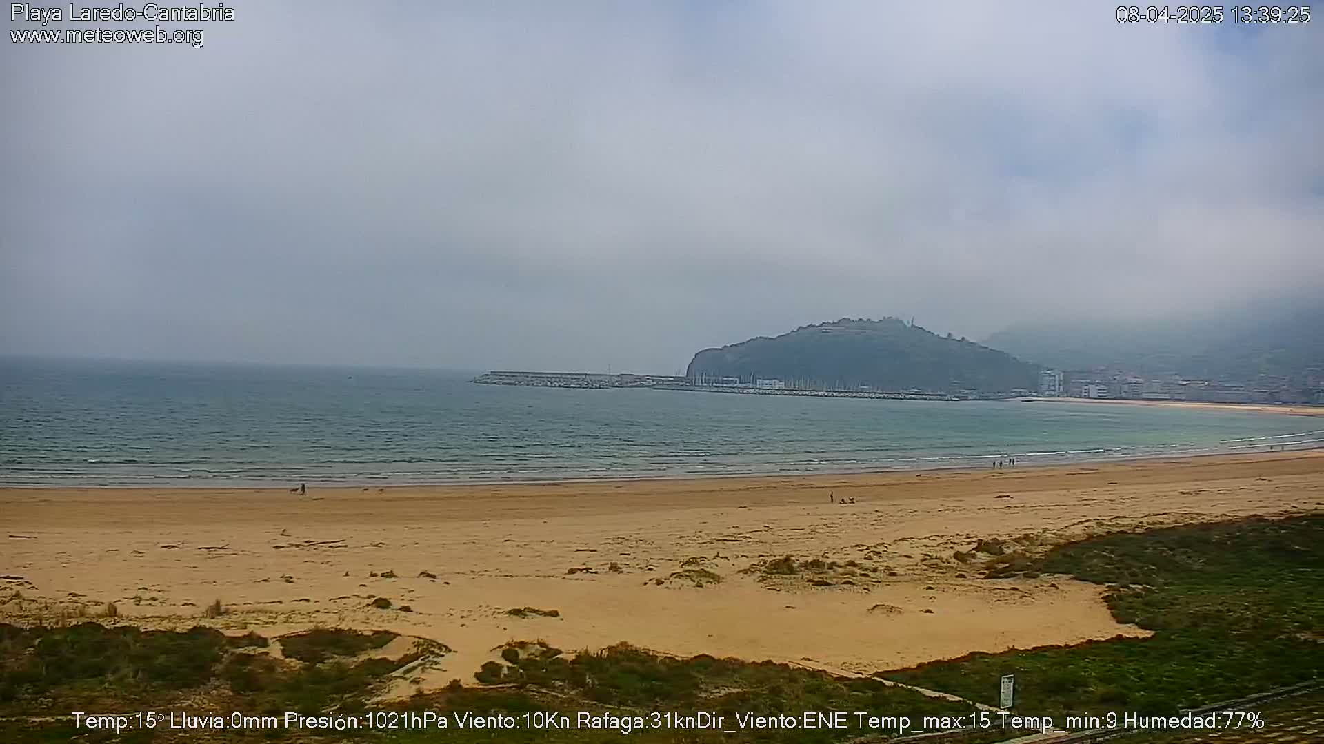 A sandy beach stretches to a calm sea under an overcast sky, with a distant hill visible.