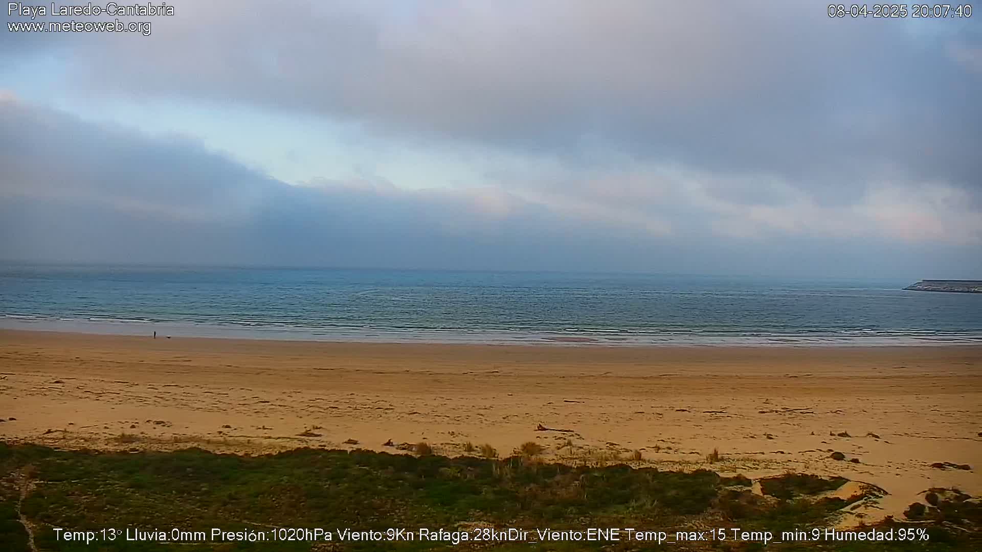 A mostly cloudy day shows a wide expanse of sandy beach and calm ocean water, with a single person visible in the distance.