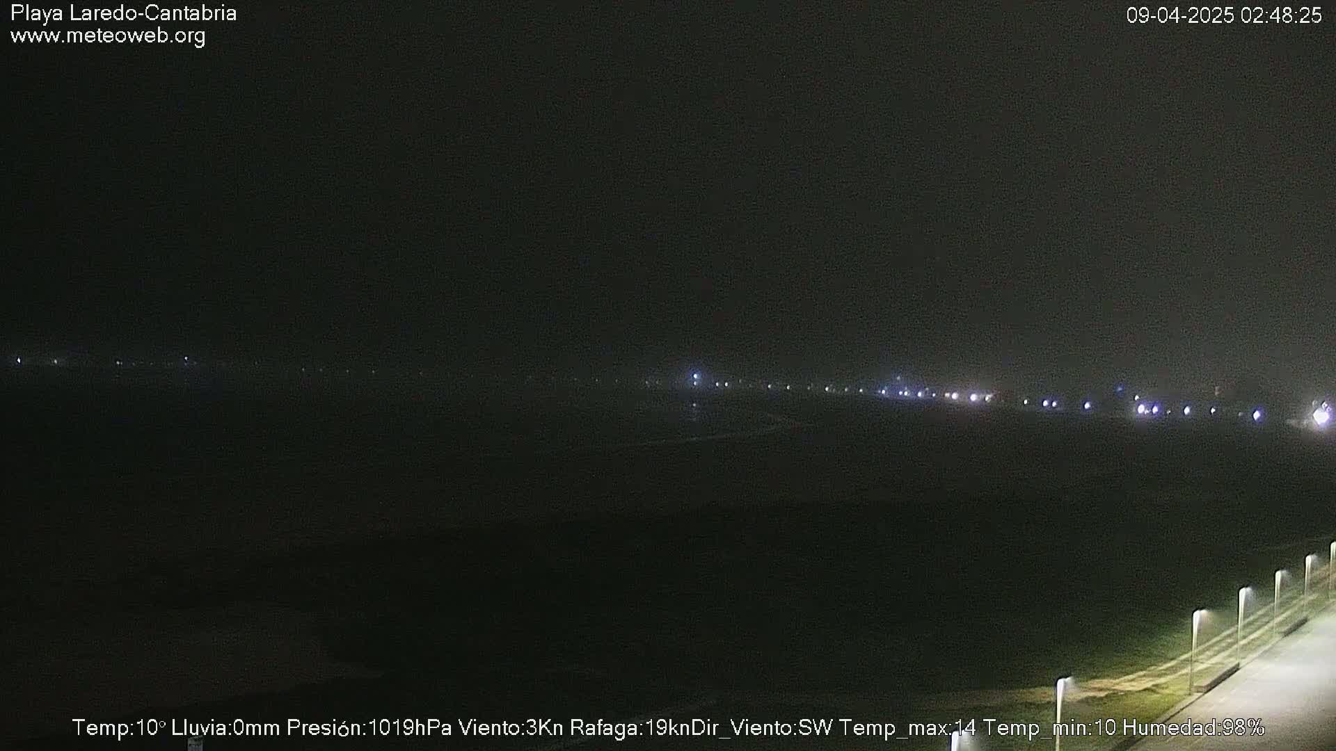 A nighttime long shot of a dimly lit coastal area with a faint line of lights in the distance under a dark, cloudy sky.