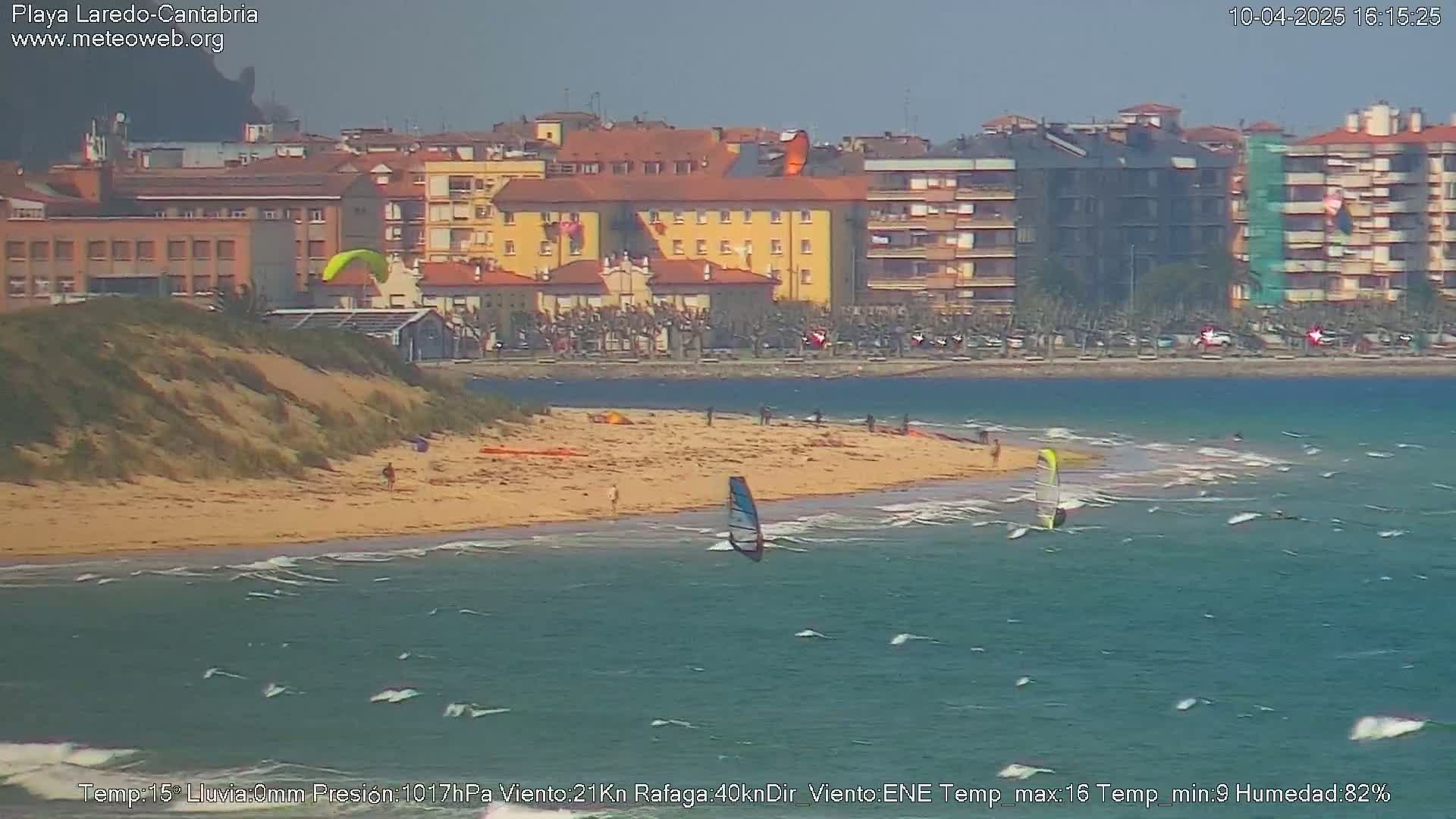 A sandy beach with several people and windsurfers is visible, with buildings and a town in the background under somewhat cloudy skies.