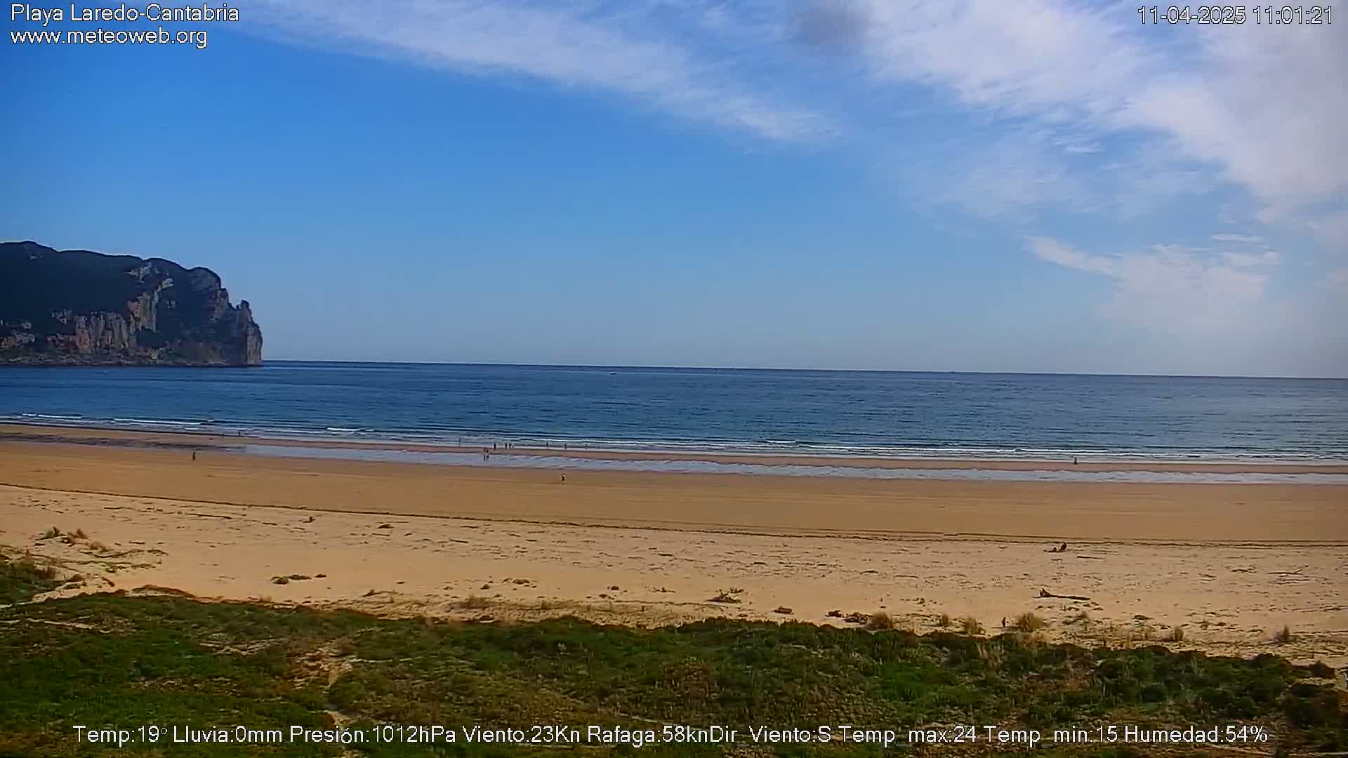 A wide sandy beach stretches to a calm ocean under a mostly clear blue sky, with a large cliff visible in the distance.