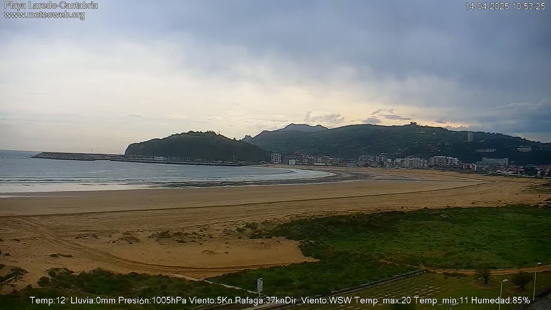 A long, sandy beach stretches out to a calm sea under a mostly cloudy sky, backed by a town nestled at the foot of rolling green hills.