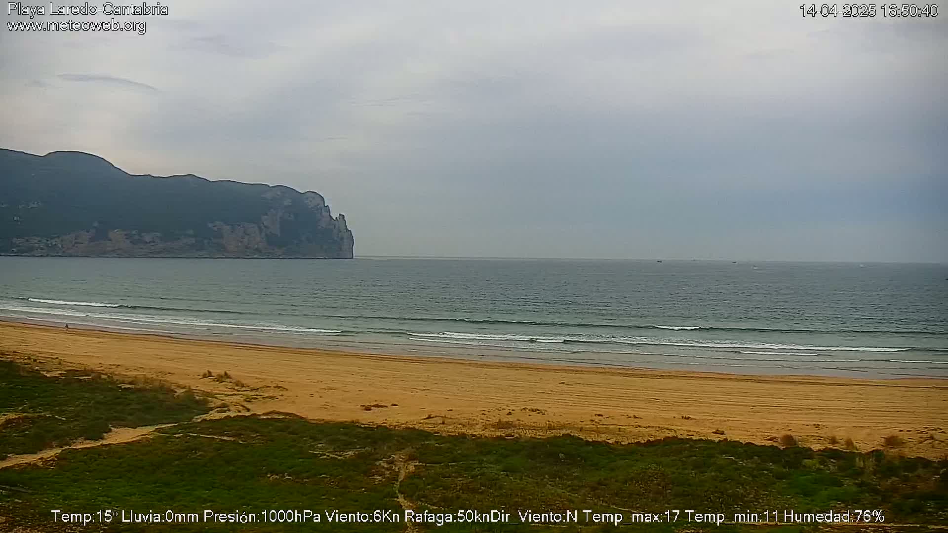 A sandy beach with gentle waves laps the shore under a cloudy sky, a distant mountain range visible across the water.