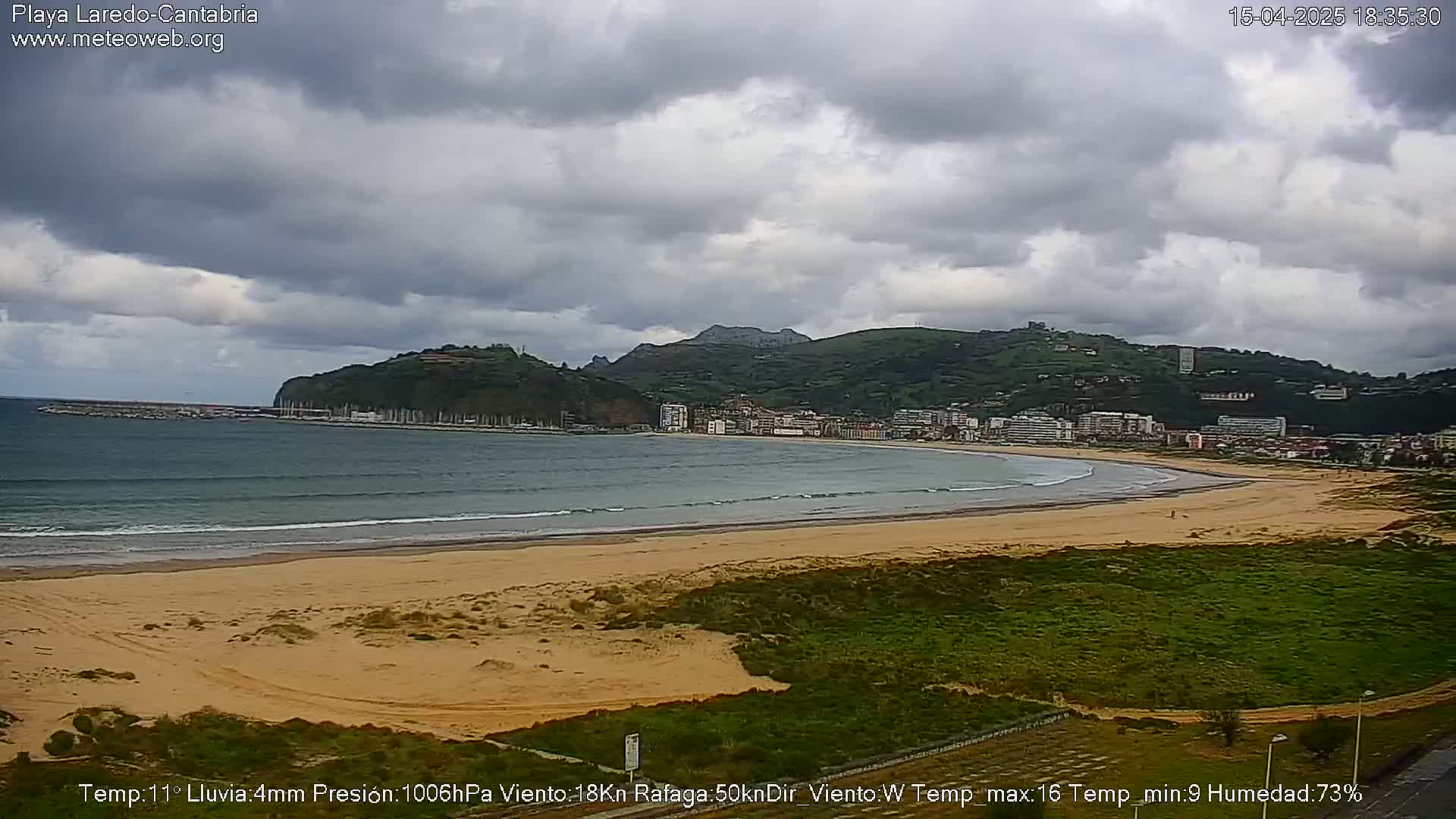 A sandy beach stretches along a bay, under a cloudy sky, with a town nestled against hills in the background.