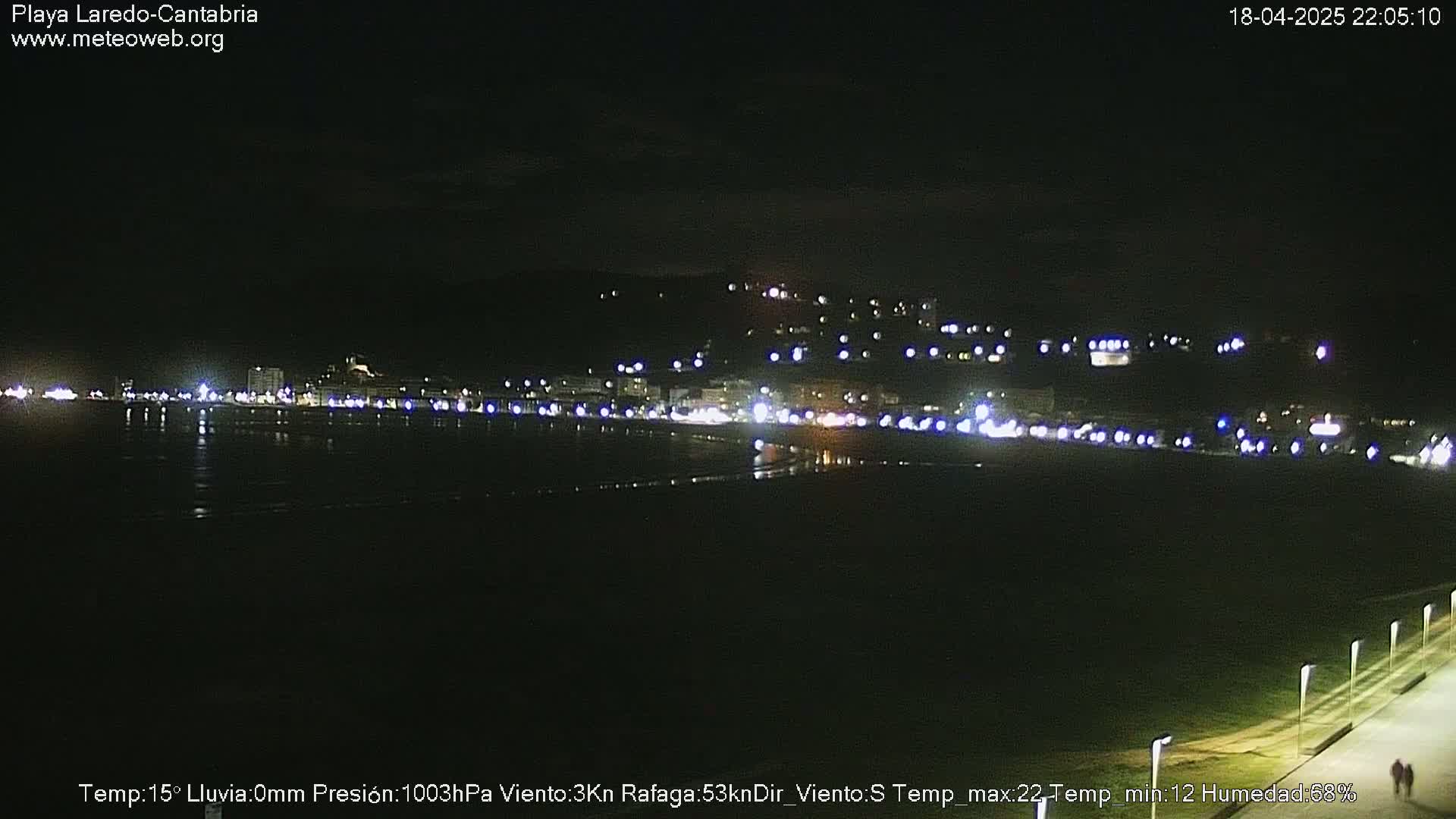 A nighttime view of a coastal town with its lights reflecting on a dark beach under a clear sky.